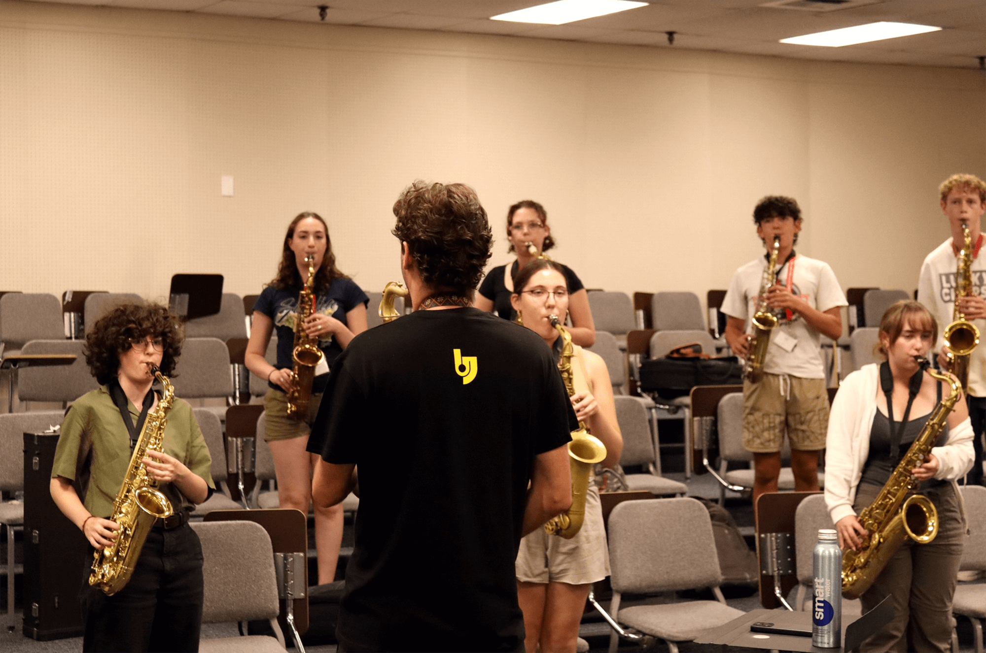 Ben Flocks conducting a group of young students playing saxophones during a practice session in a classroom or rehearsal room.