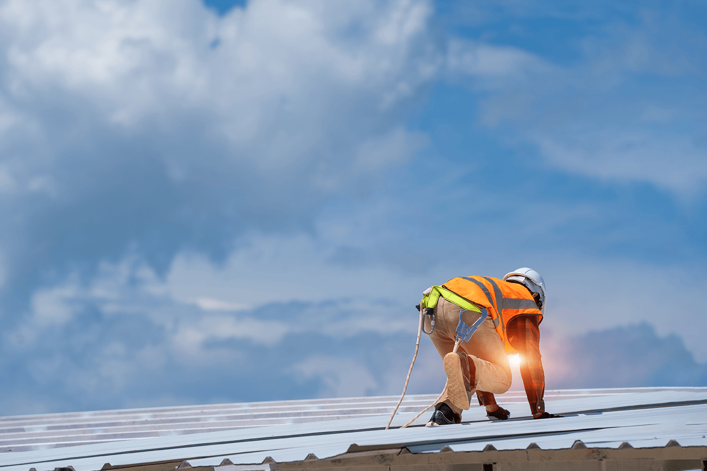 A construction worker wearing an orange safety vest and helmet, crouched on a roof, working on installing solar panels with a bright sky and clouds in the background.