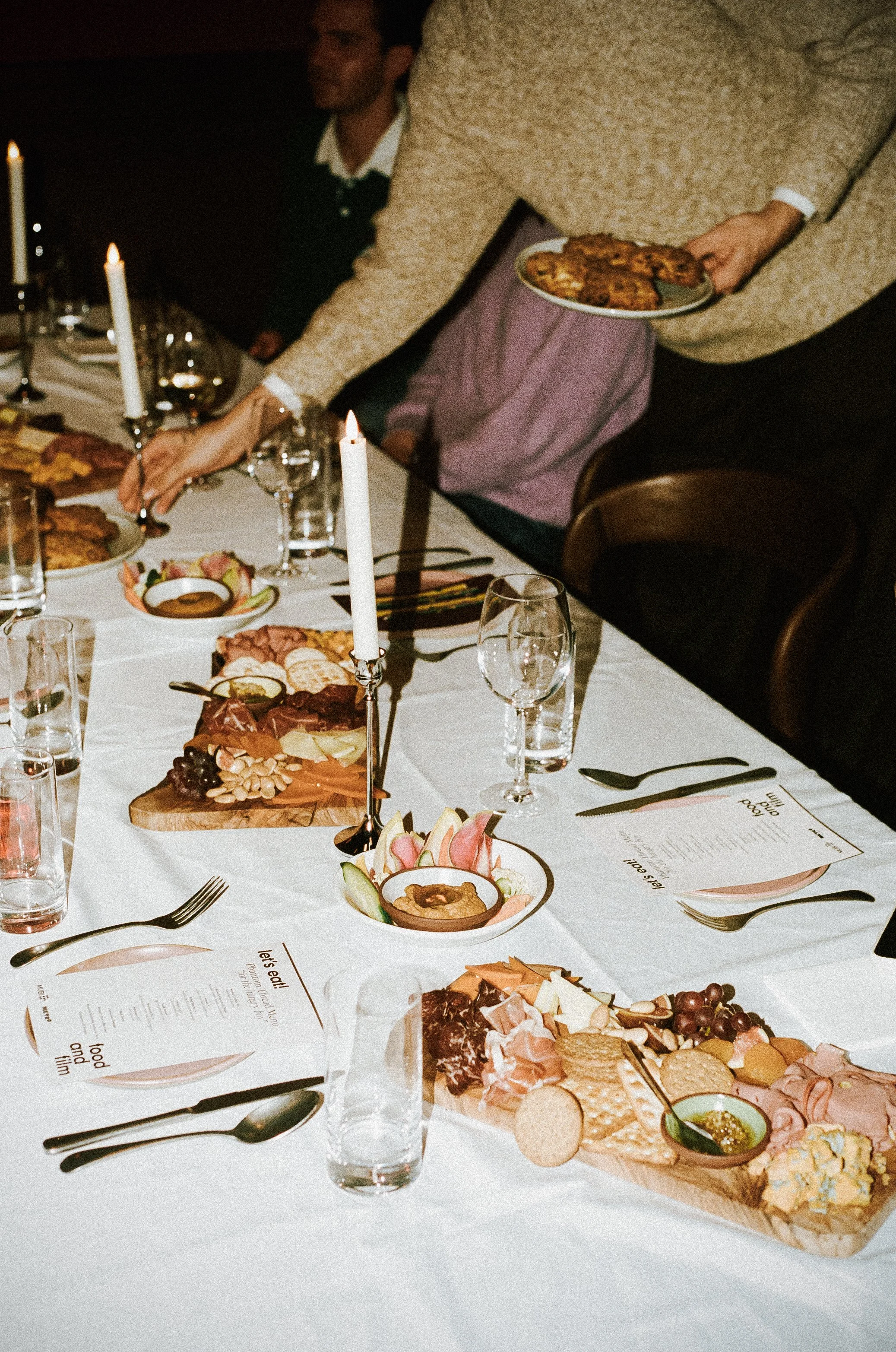 A dining table set for a meal with appetizers, cheeses, and charcuterie, illuminated by candles, with a person serving food on a plate.