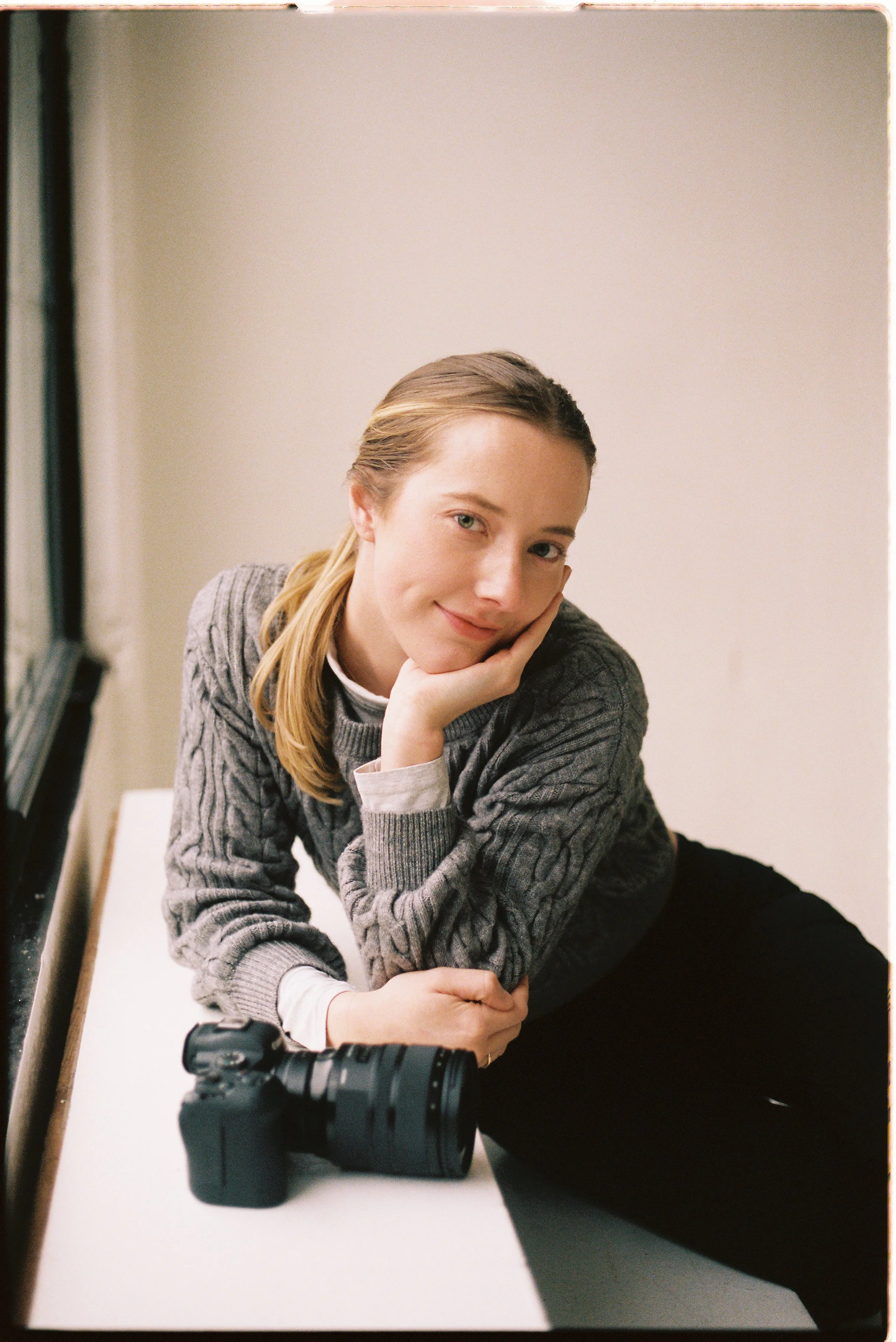 A young woman with blonde hair, wearing a gray cable-knit sweater, is sitting on a table with a camera nearby, resting her chin on her hand and smiling at the camera.