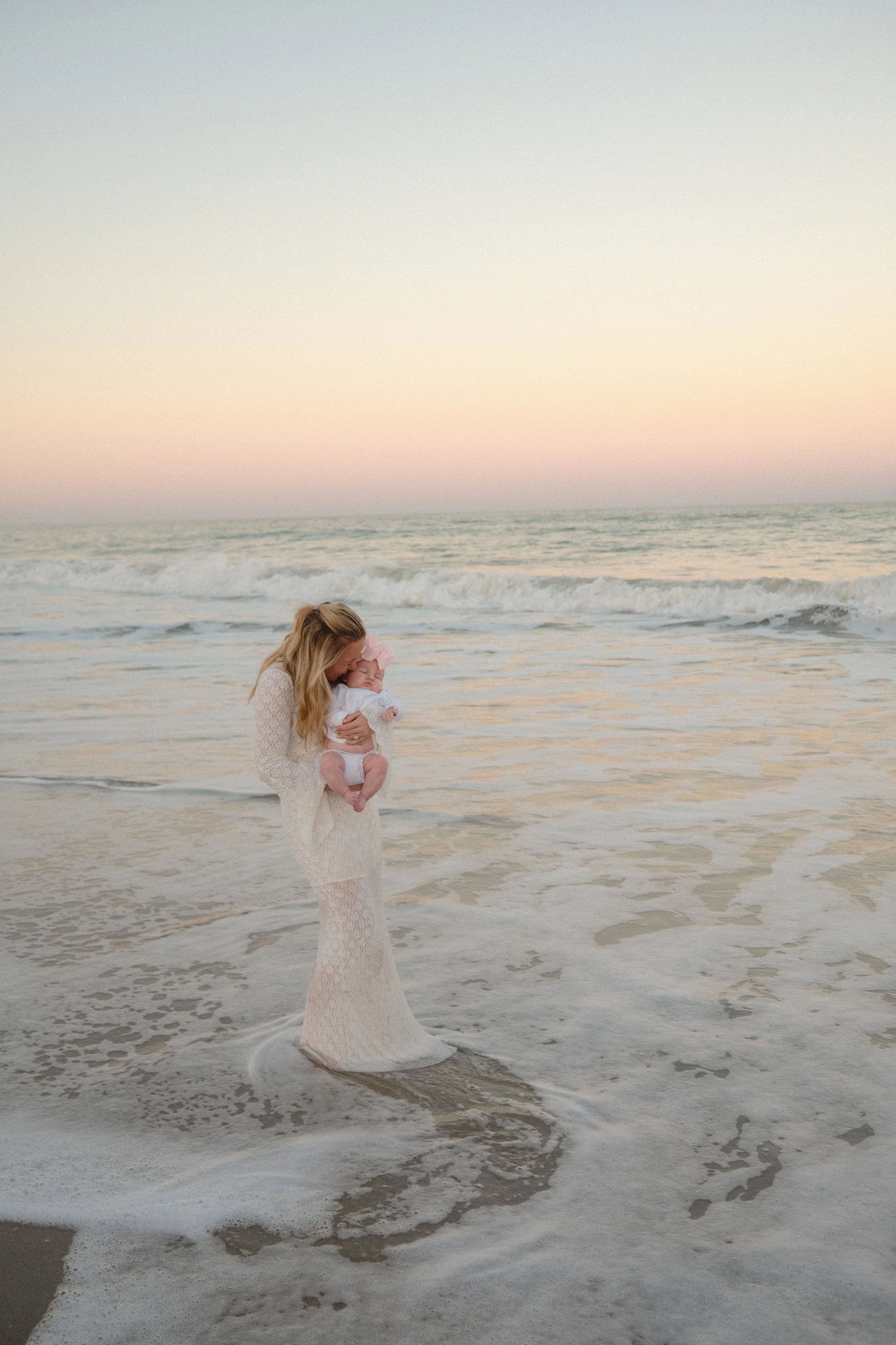A woman holding a baby at the beach during sunset, with gentle waves and a pastel-colored sky in the background.