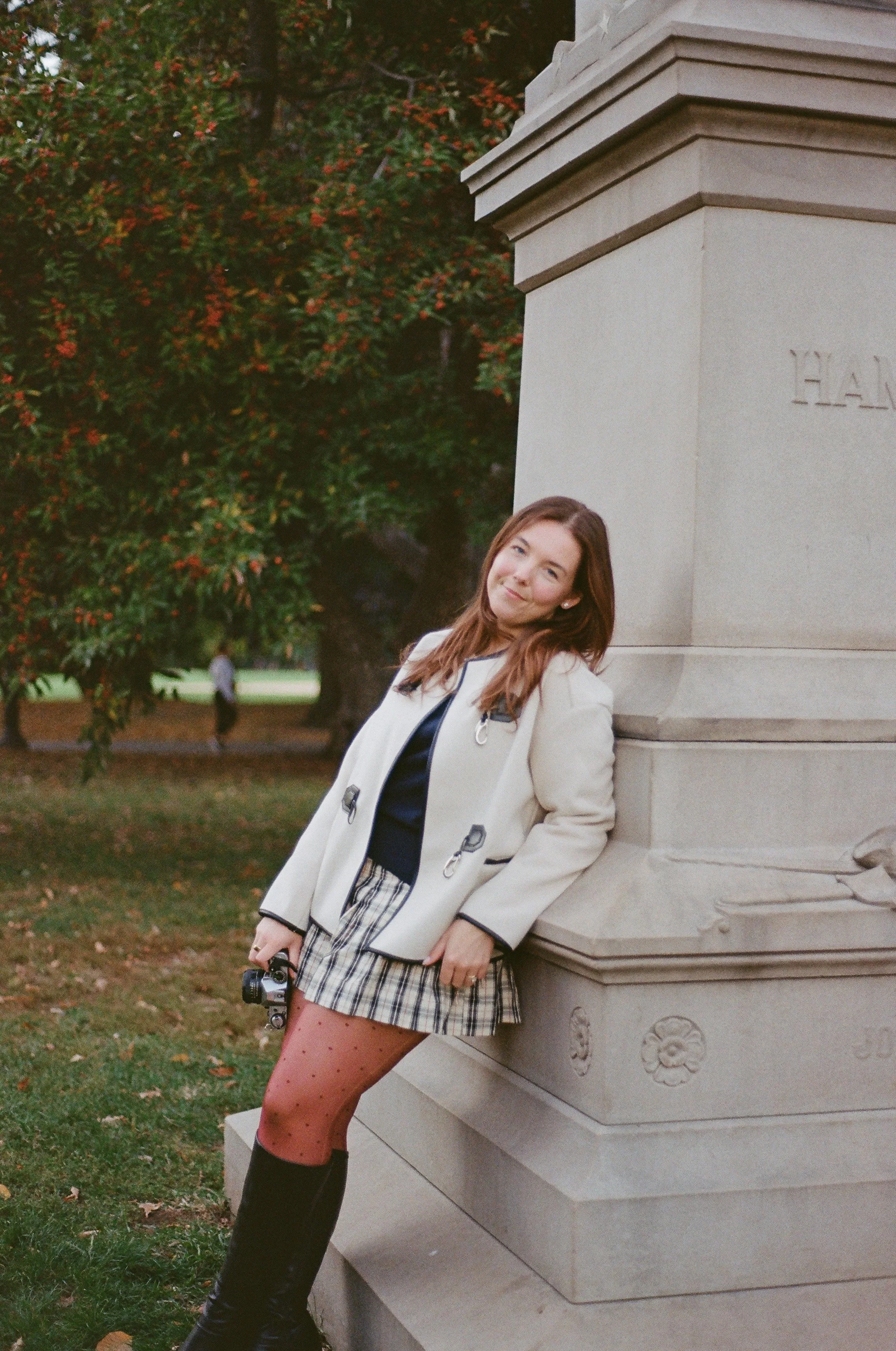A young woman with long brown hair leaning against a stone monument in a park, holding a small camera, wearing a cream blazer, black top, plaid skirt, pink polka dot tights, and black knee-high boots, with red and green trees in the background.