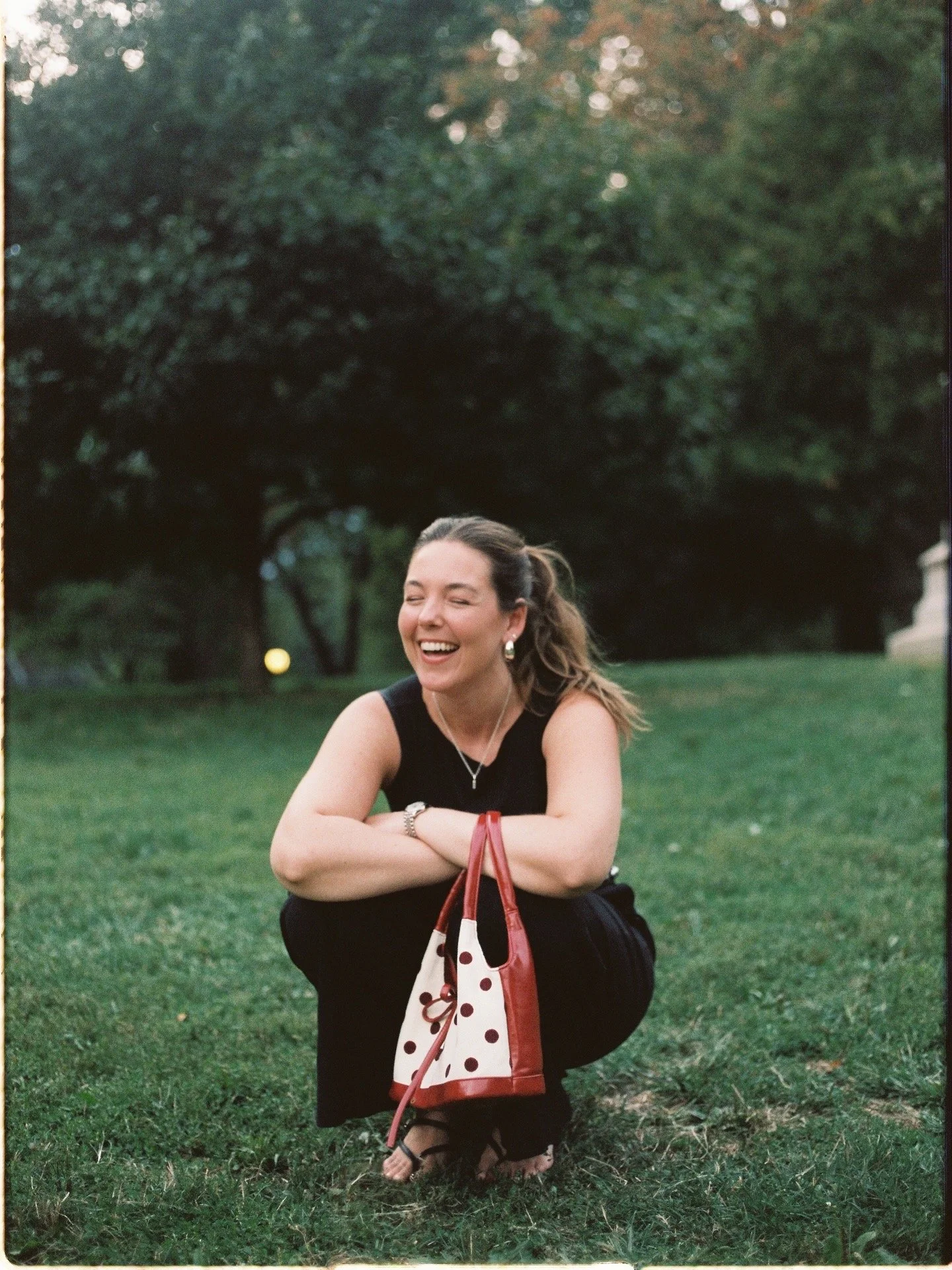 Woman with brown hair in a ponytail, smiling and squatting on grass in a park, holding a red and white polka dot handbag, wearing a black sleeveless top, black pants, and sandals, with trees and a building in the background.