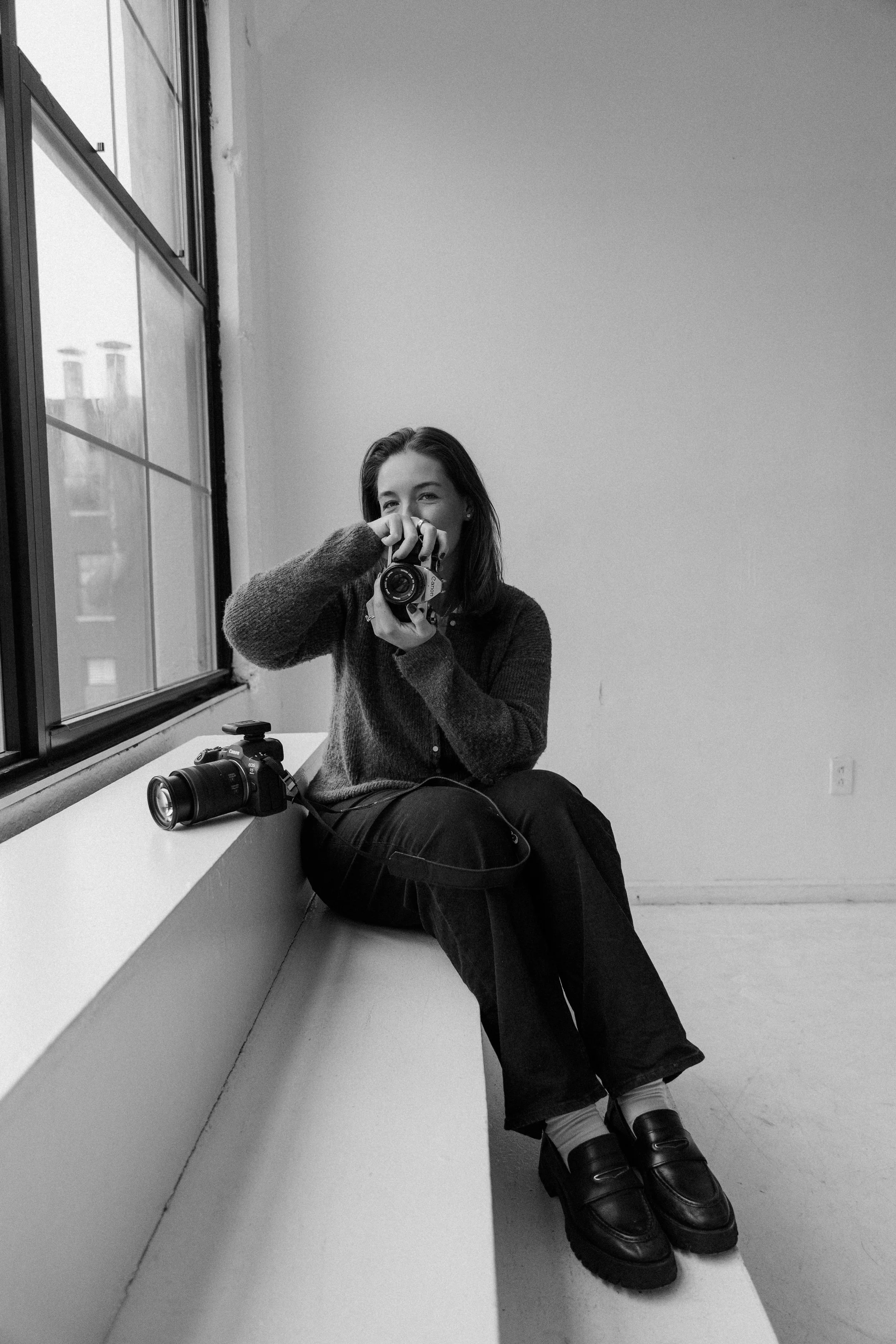 A young woman sitting on a windowsill in a room, smiling and holding a camera to her eye, with another camera placed beside her. The room has a large window with a cityscape view outside.