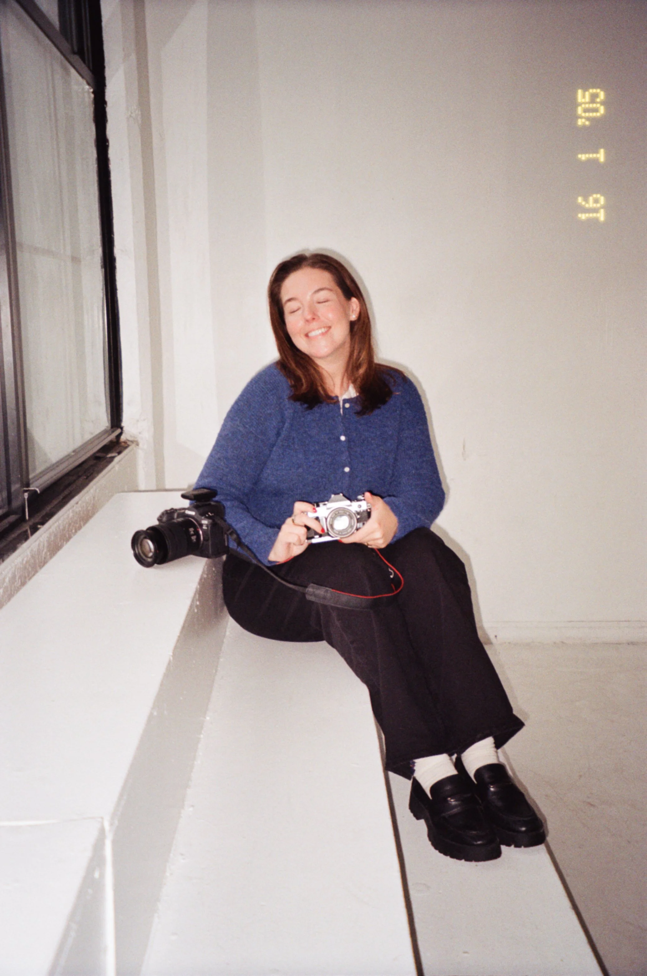 A young woman with red hair, wearing a blue sweater, black pants, and black shoes, sitting on a white bench in a room with white walls and a window, holding a camera while smiling with eyes closed.