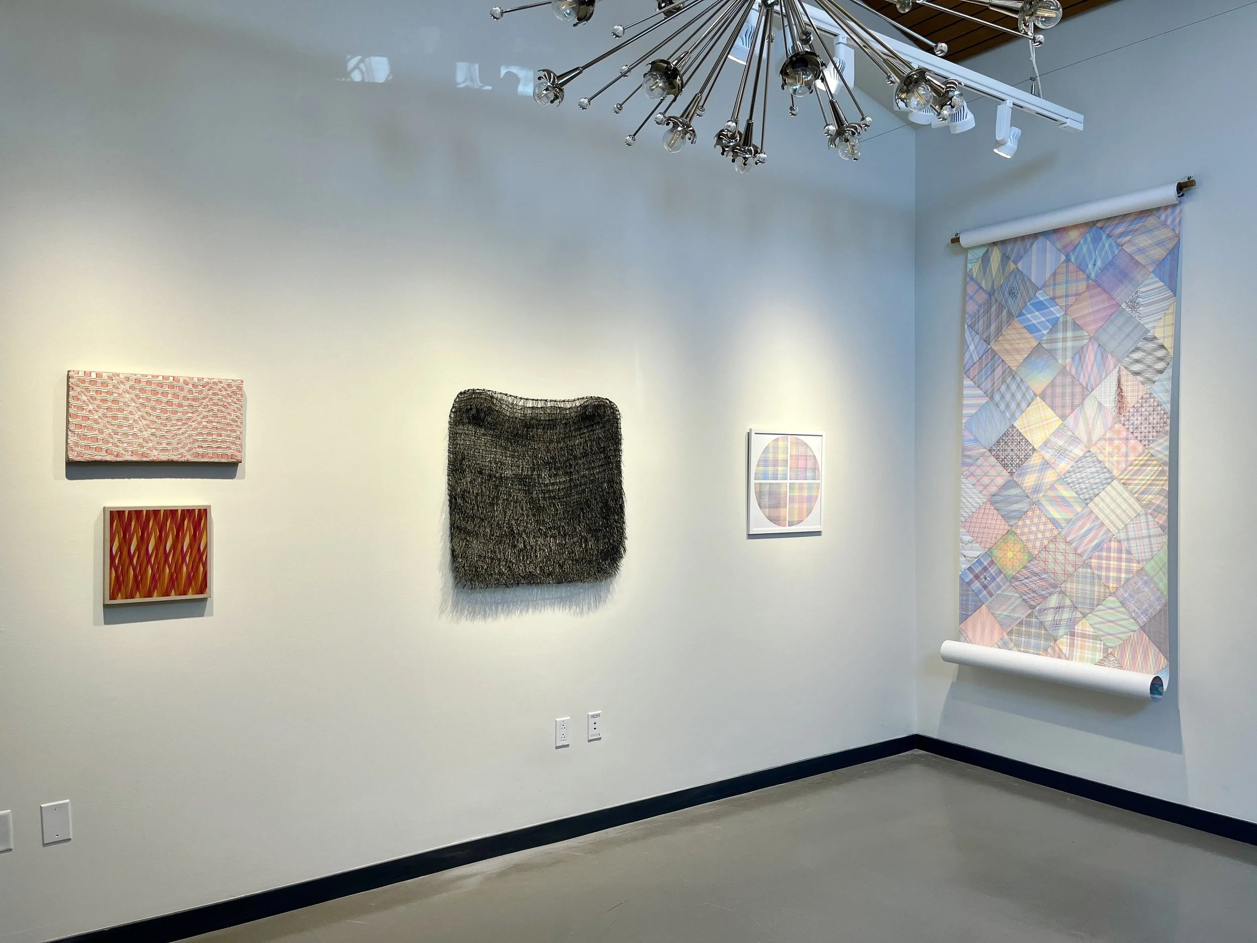Art gallery wall with abstract and textured artwork, including a woven black piece, a small red and orange pattern, and a geometric pastel composition, with a contemporary light fixture hanging from the ceiling and a colorful patchwork textile scroll on the right side.