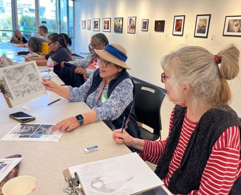 Group of older women sitting at a table in an art gallery, looking at artwork and sketches, with others engaging in similar activities in the background.