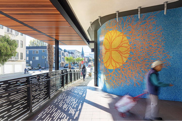 A sidewalk covered porch with a large colorful mural of a tree on a blue wall, overlooking a city street with pedestrians, buildings, and palm trees.
