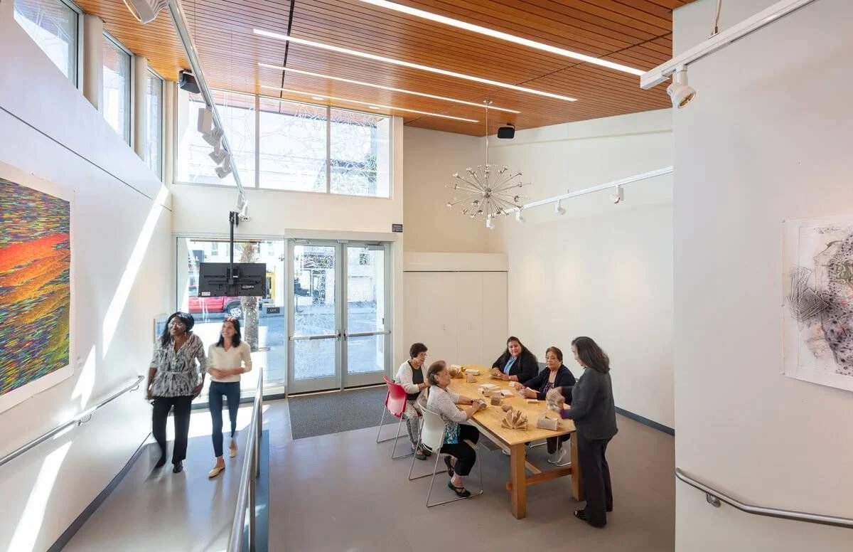 People sitting at a table in a bright art gallery or studio, engaging in a workshop or discussion, with artwork on the walls and large windows allowing natural light.