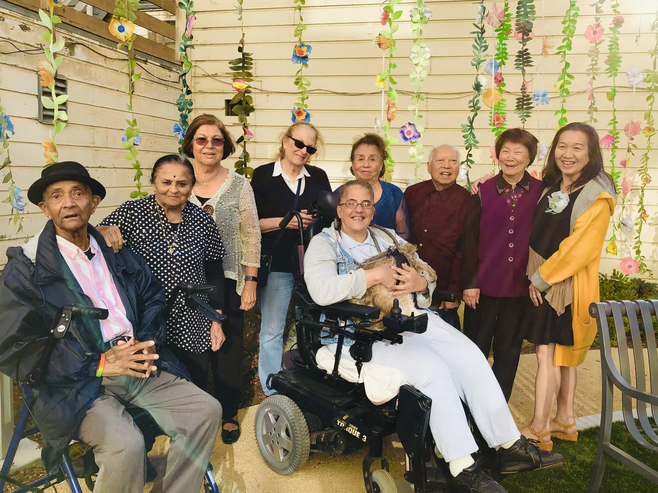 Group of diverse elderly people and caregivers smiling outdoors with colorful paper flower decorations hanging behind them.
