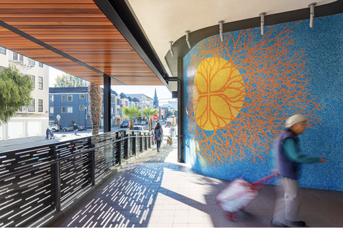Urban outdoor walkway with colorful mural of a yellow sun and orange tree on blue wall, people walking, and residential buildings in the background.