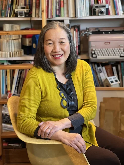 A woman with shoulder-length gray hair sitting in a yellow armchair, smiling at the camera, in a room with bookshelves filled with books and vintage cameras, and a typewriter in the background.