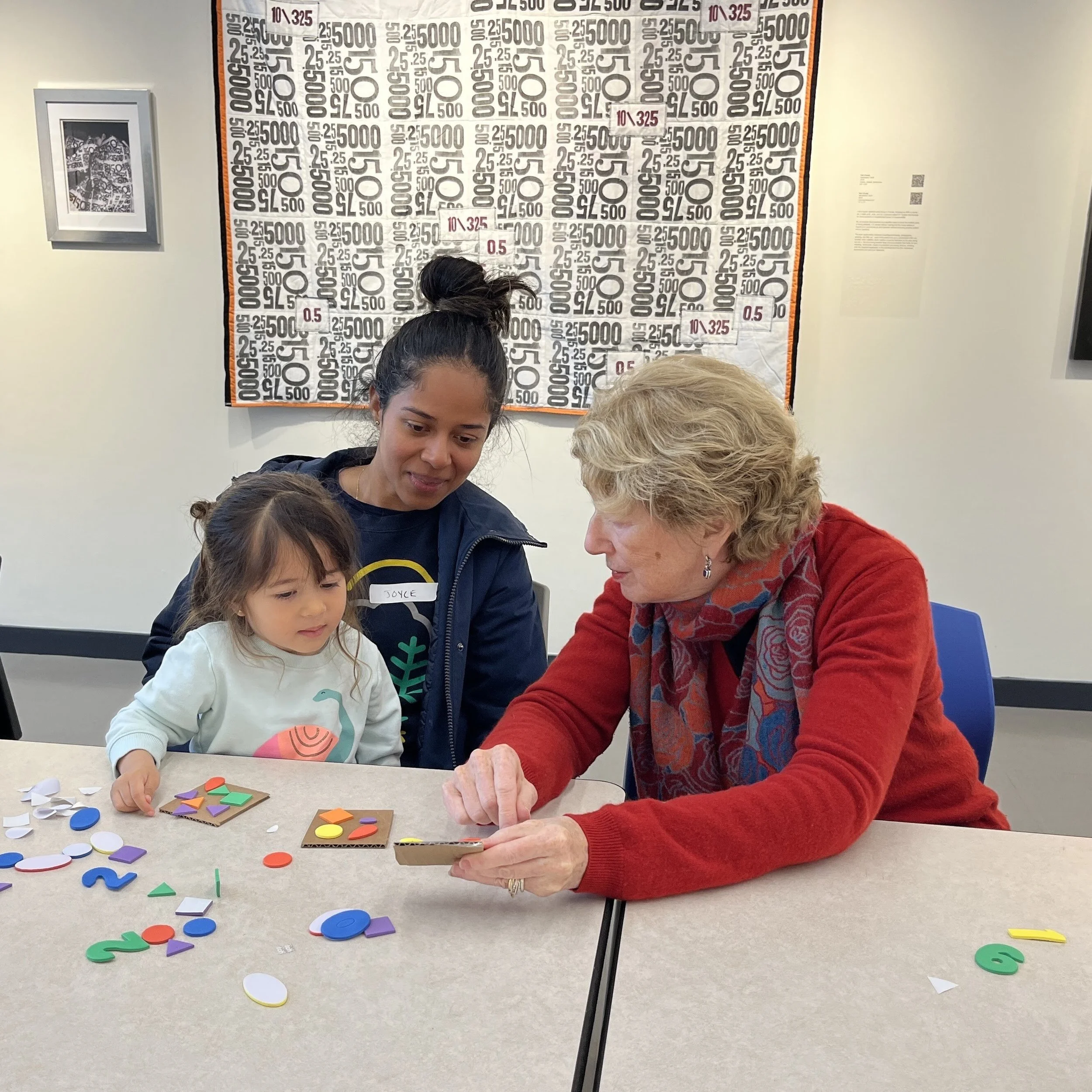 Three people, including a young girl, an older woman, and a younger woman, are sitting at a table playing a colorful shape and number puzzle game. A large piece of artwork made from black, white, and purple numbers and symbols is on the wall behind them.
