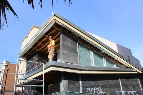 A construction worker standing on a balcony of a modern house under construction, with scaffolding in front, against a clear blue sky.
