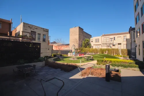 Courtyard garden with benches, trees, and shrubbery, surrounded by apartment buildings under a clear blue sky.