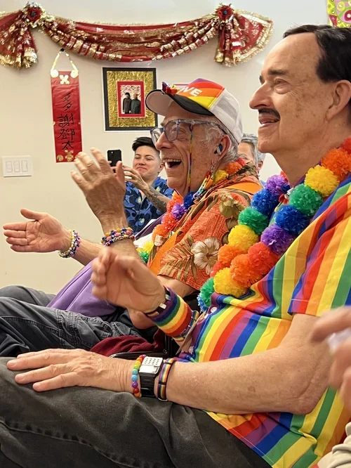 People dressed in colorful, festive clothing with rainbow leis, celebrating at a party or gathering, with decorations on the wall.