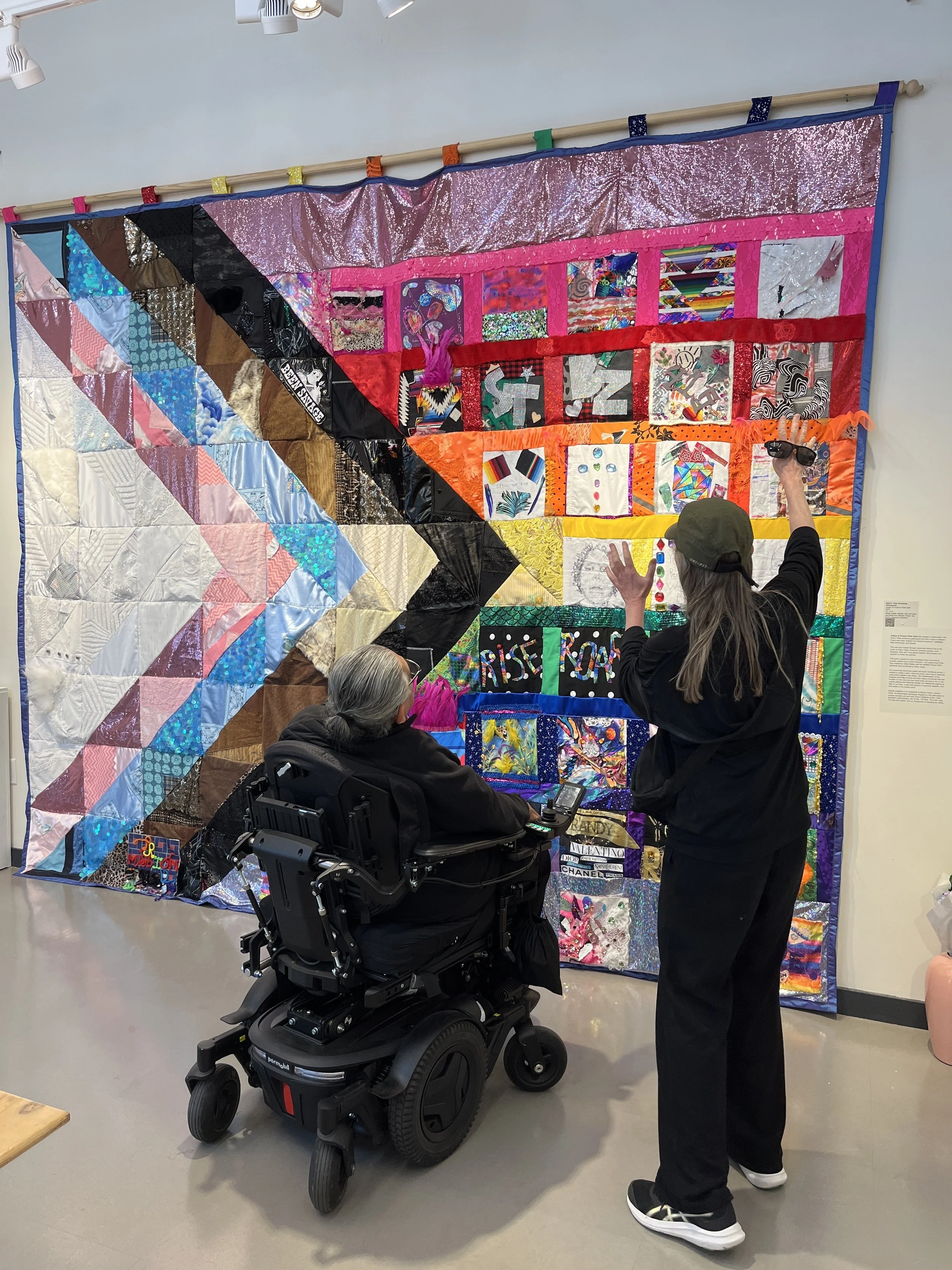 A person in a wheelchair and a woman are looking at a large, colorful quilt displayed on a wall. The quilt features various patterns, textures, and colors, with several sections bordered by pink and orange fabrics.