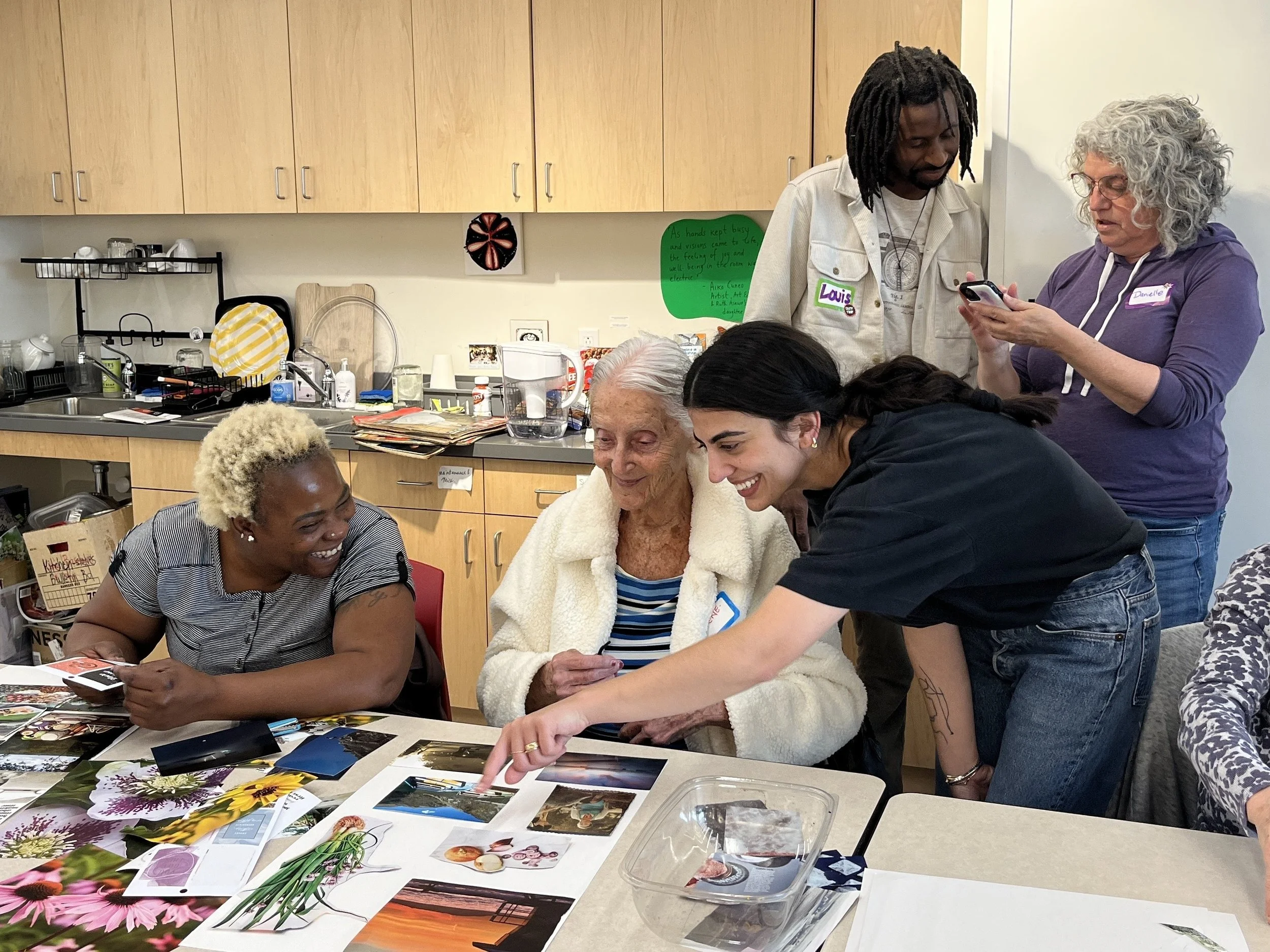 Group of diverse people gathered around a table looking at photographs and artwork, engaging in an activity together in a room with kitchen cabinets and decorative wall art.