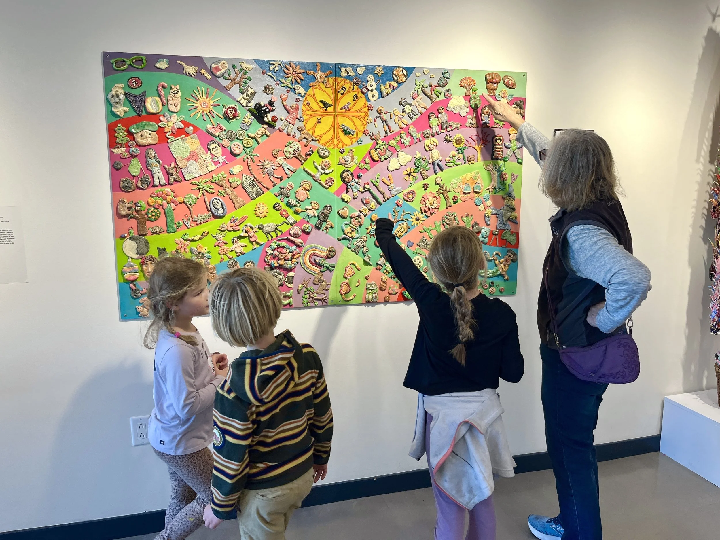 Children and an adult observing a colorful art display made of various clay figures arranged in a pattern on a wall.