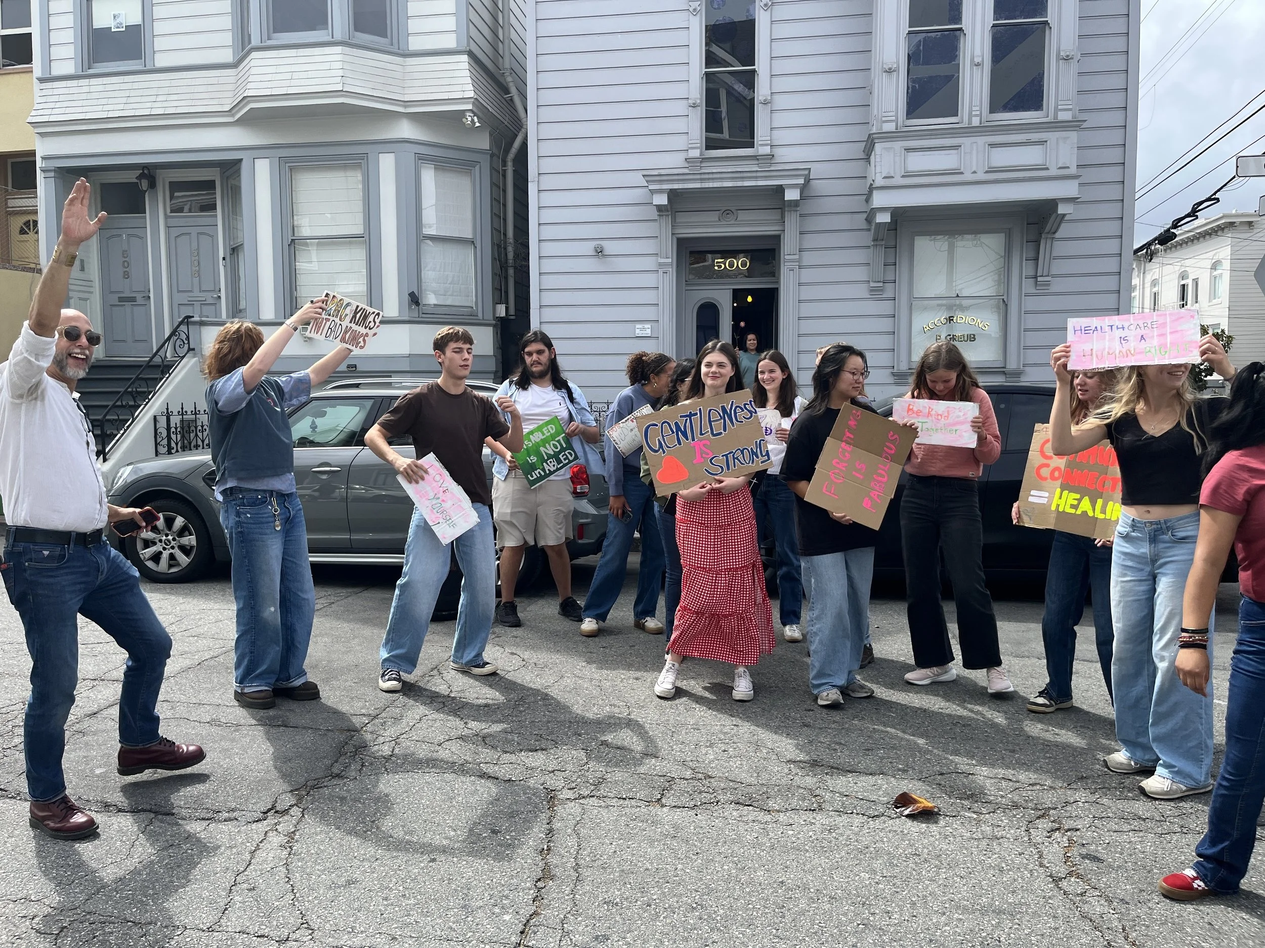 A group of people protesting on a sidewalk in front of a gray house, holding signs with messages about healthcare and kindness, while some people are smiling and waving.