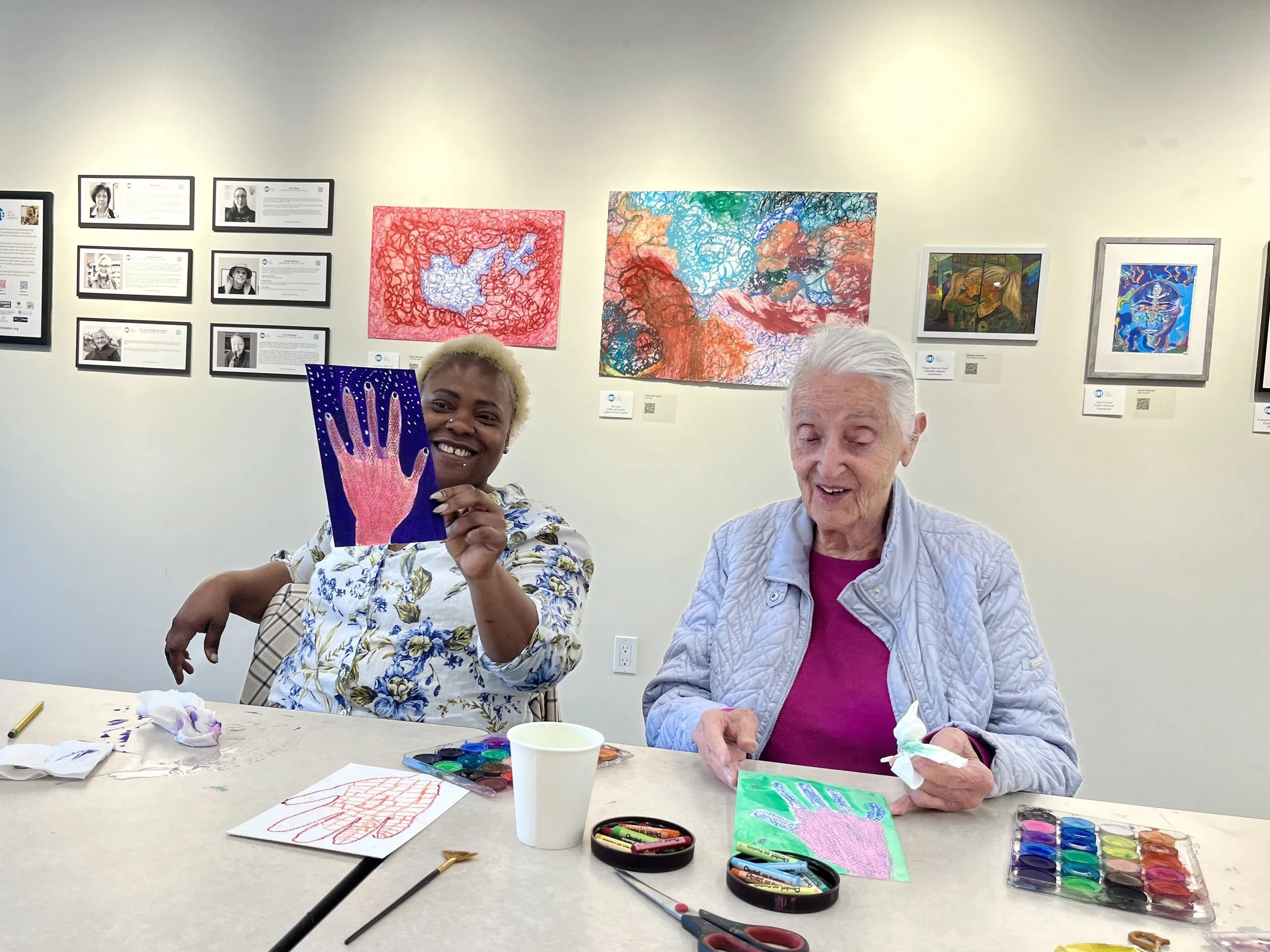 Two women seated at a table creating artwork with paints, brushes, and paper, in an art gallery with framed art on the walls.