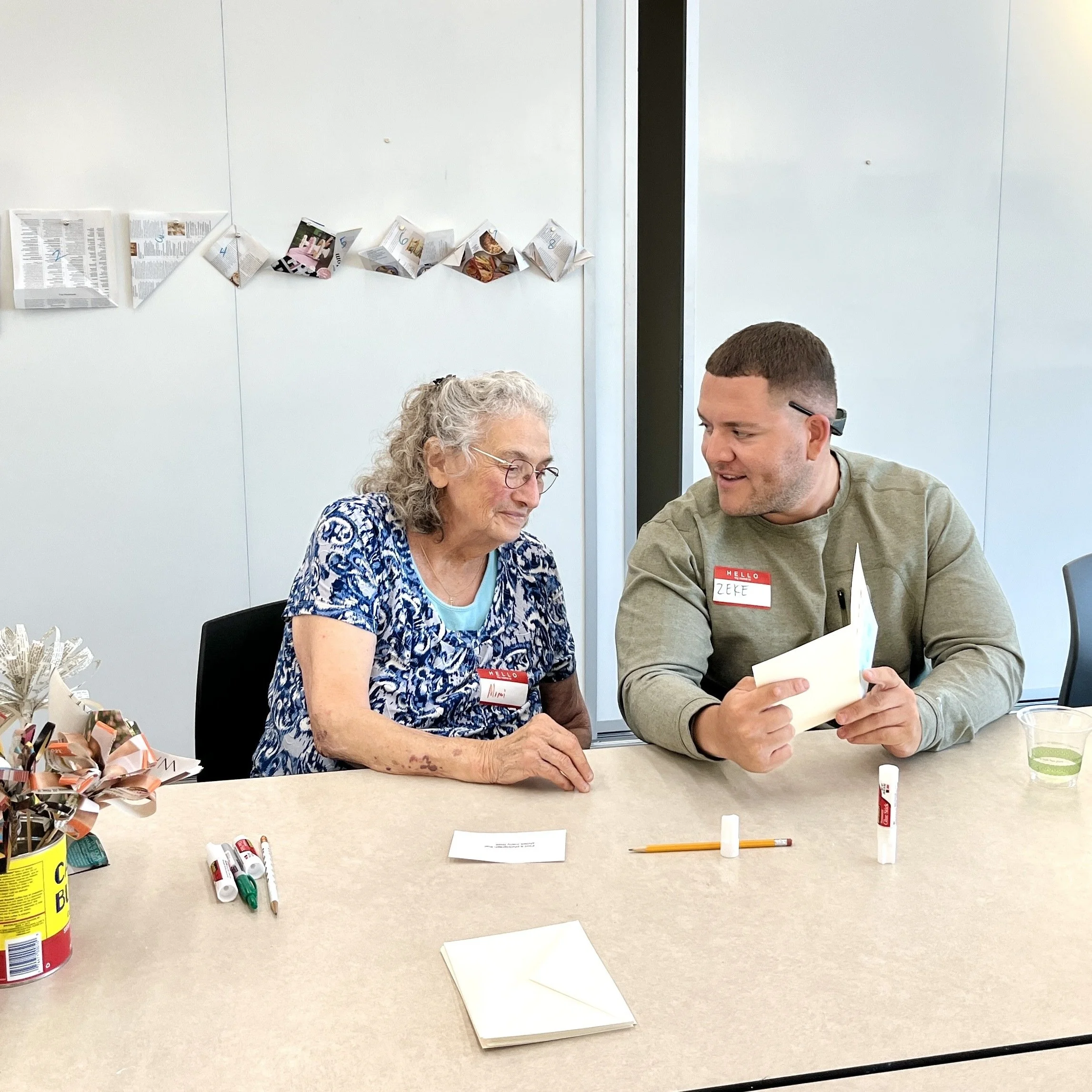 Two people, a senior woman and a young man, sitting at a table engaged in a conversation. The woman has white curly hair, glasses, and is wearing a blue patterned blouse. The man has short brown hair and is wearing a green shirt with a red name tag. On the table, there are white papers, colored markers, and a disposable cup. Behind them, a plain white wall with a string of folded paper decorations and a visible door frame.