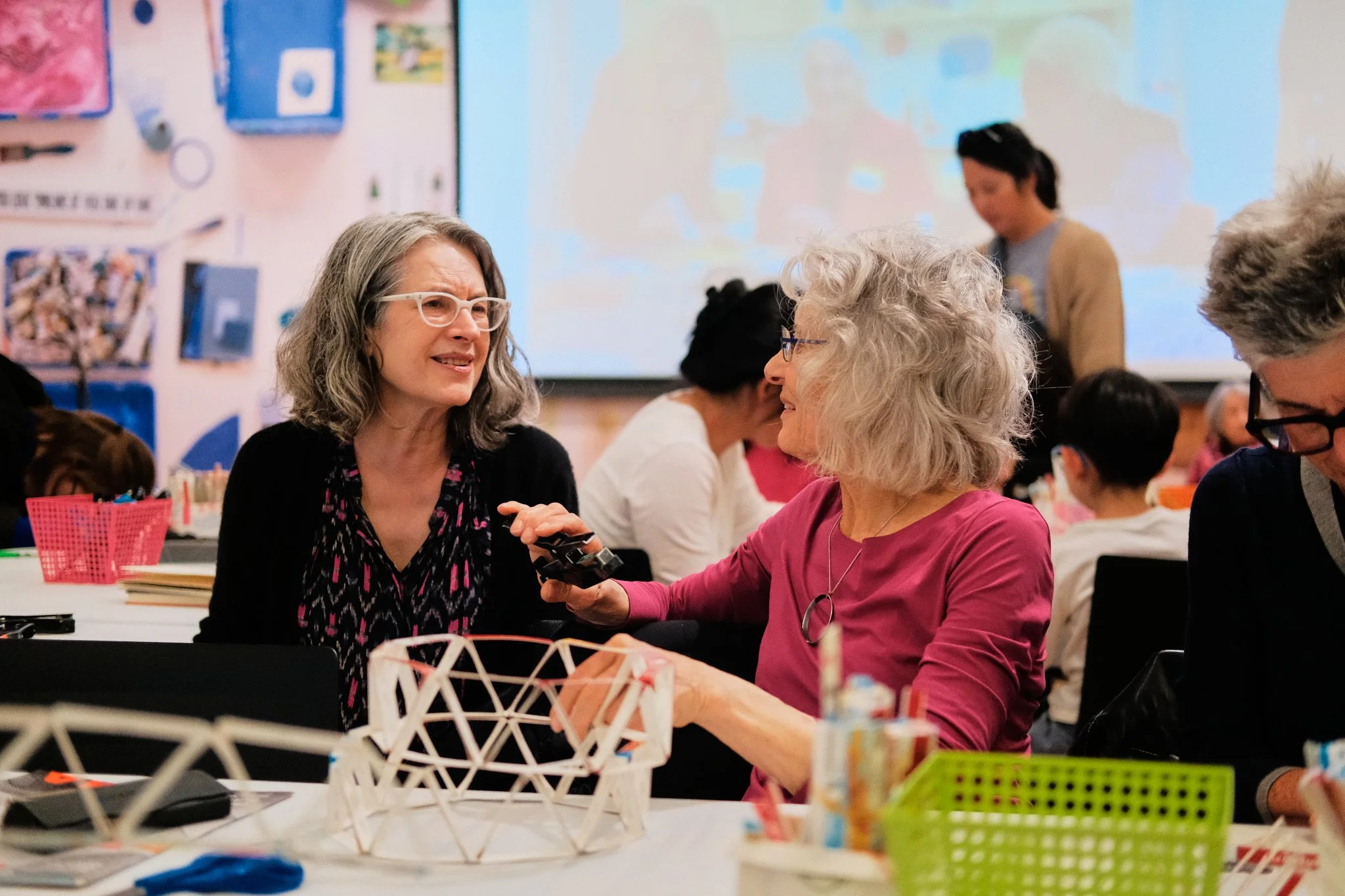 Two women are engaged in conversation at a table during a workshop or event, with other people in the background and educational displays on the walls.