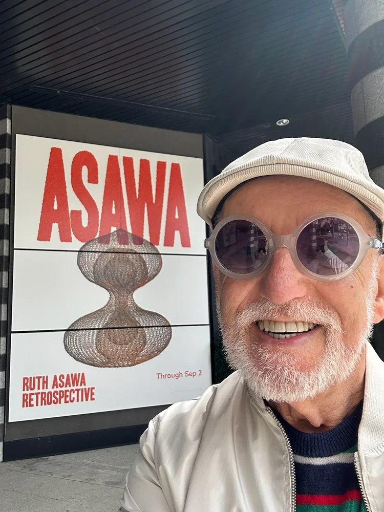 A smiling man with white hair, beard, and sunglasses taking a selfie outside an art exhibit. The display behind him reads 'Ruth Asawa Retrospective' and features a wire sculpture, with the name 'Asawa' in large red letters.