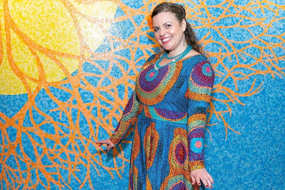 Woman in a colorful, patterned dress standing in front of a vibrant mosaic wall with orange and blue motifs.