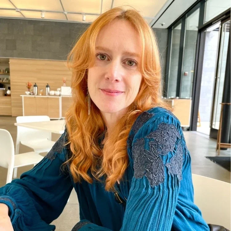 Red-haired woman sitting at a table in a modern cafe or restaurant, smiling at the camera, with large glass windows behind her.