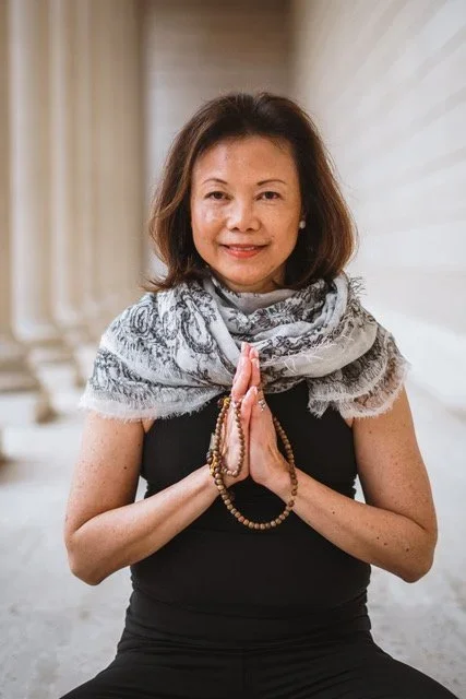 A woman sitting with hands pressed together in a prayer position, holding a beaded necklace, smiling, in a corridor with columns.