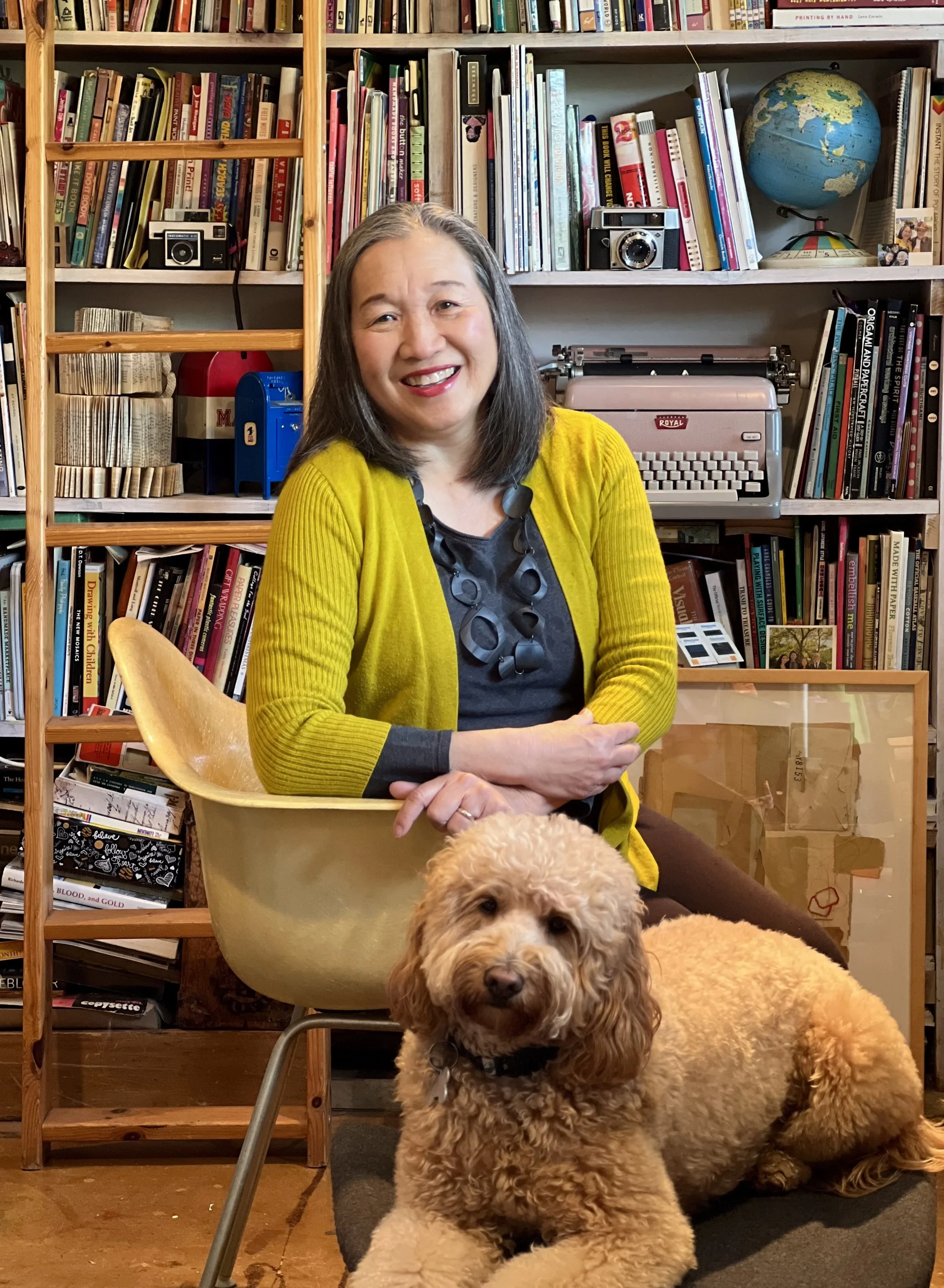 A woman with gray hair wearing a yellow cardigan sitting on a chair with a large fluffy dog in the foreground and a smaller similarly fluffy dog on a mat in front. The background features a bookshelf filled with books, a vintage typewriter, and a globe.