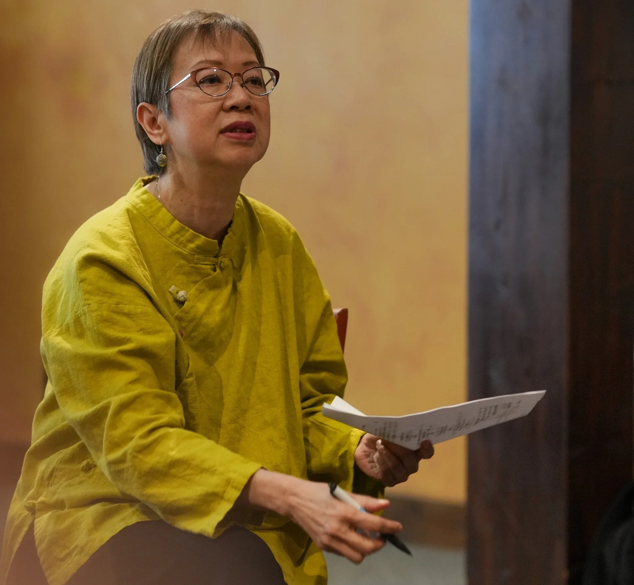 A woman with short gray hair and glasses wearing a yellow top and earrings standing while holding papers and a pen, at an indoor event or presentation.