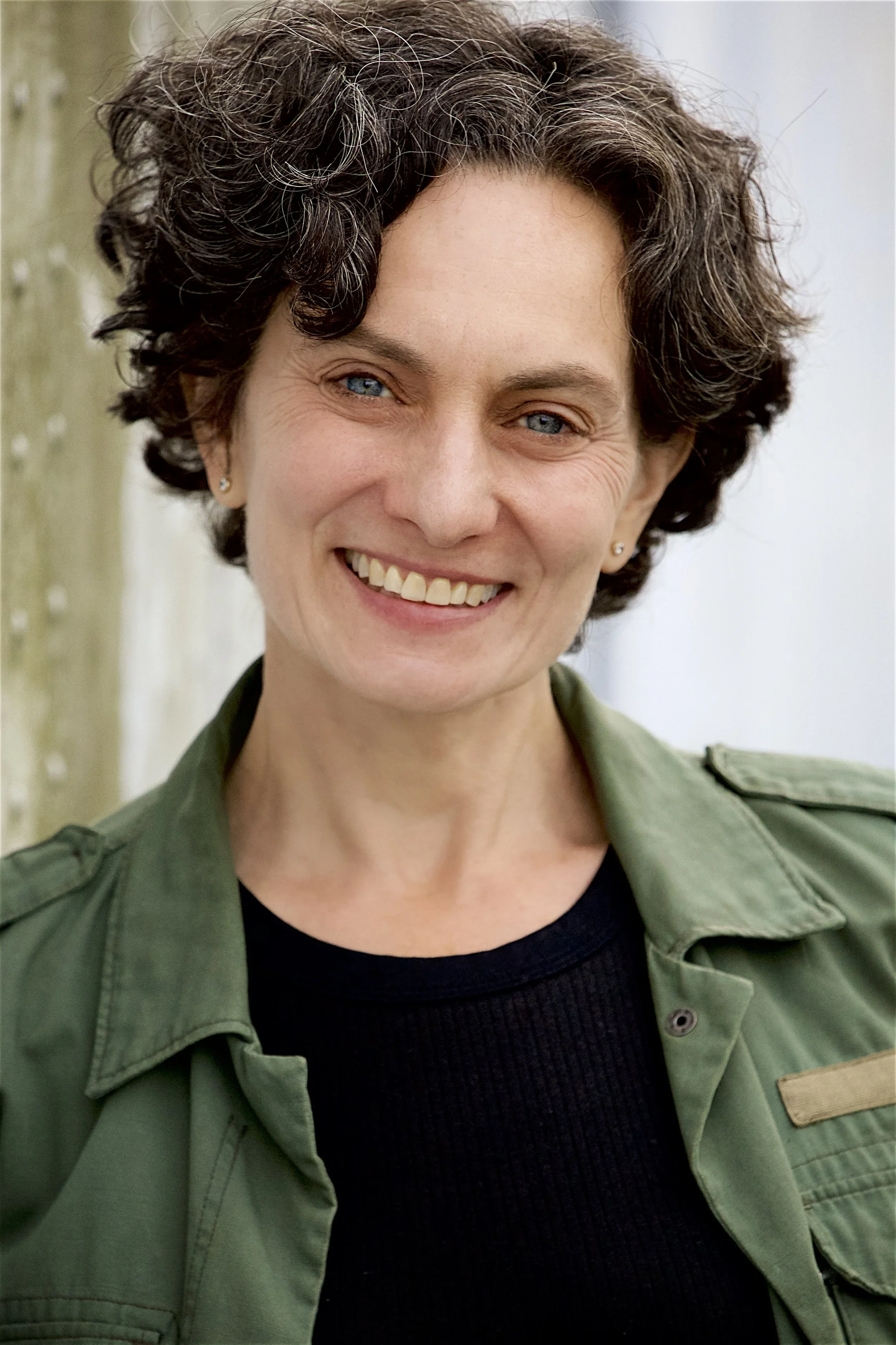 Close-up of a woman with short, curly brown hair, blue eyes, wearing earrings, a green military-style jacket, and a black shirt, smiling outdoors near a wooden post.