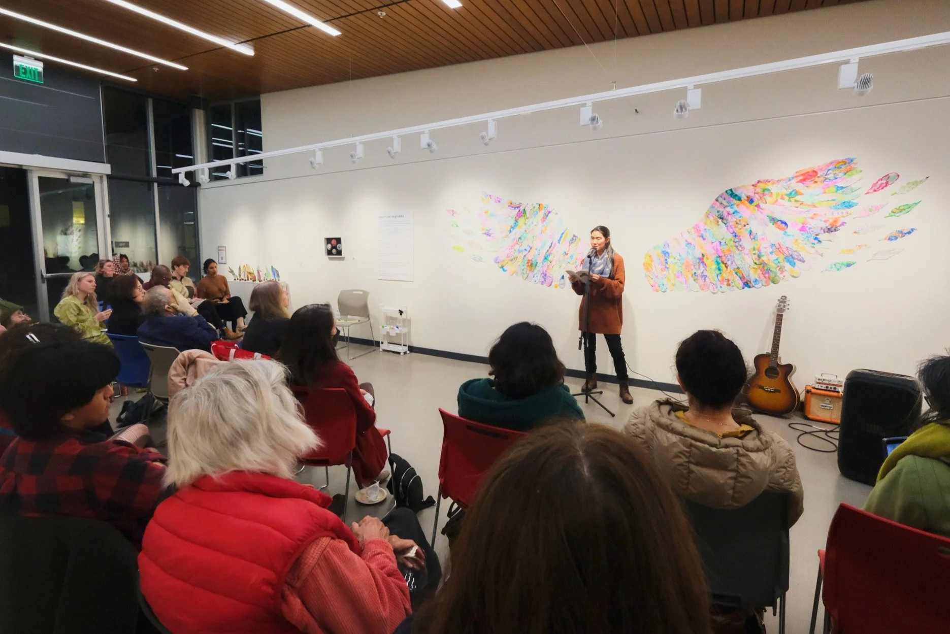 A woman is performing or speaking on a small stage with a microphone in a gallery or art space, with colorful angel wing artwork on the wall behind her. People are seated and watching the performance, and there is a guitar and amplifier on the stage.