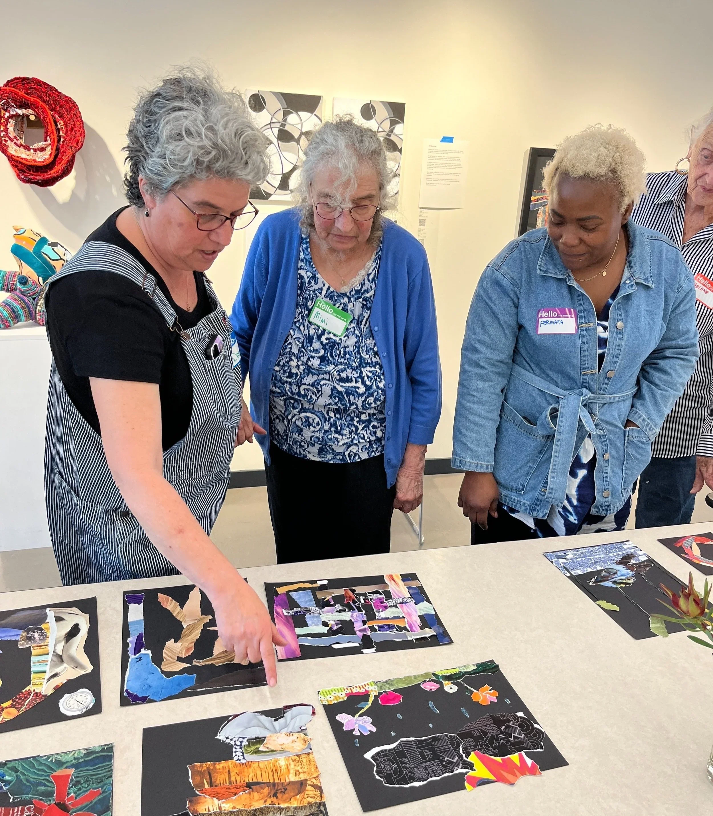 Group of women looking at and pointing to colorful paper collage art on tables in an art gallery.