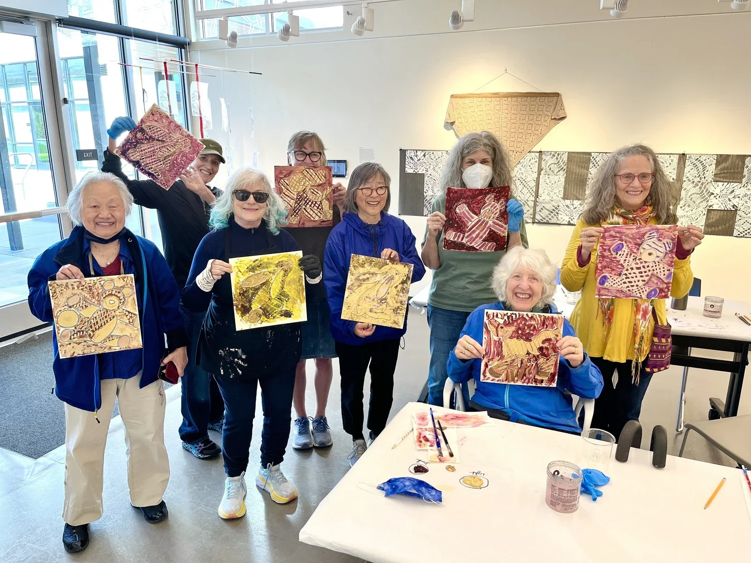 Group of eight women in an art studio holding up colorful, abstract paintings they created, smiling and showing their artwork.