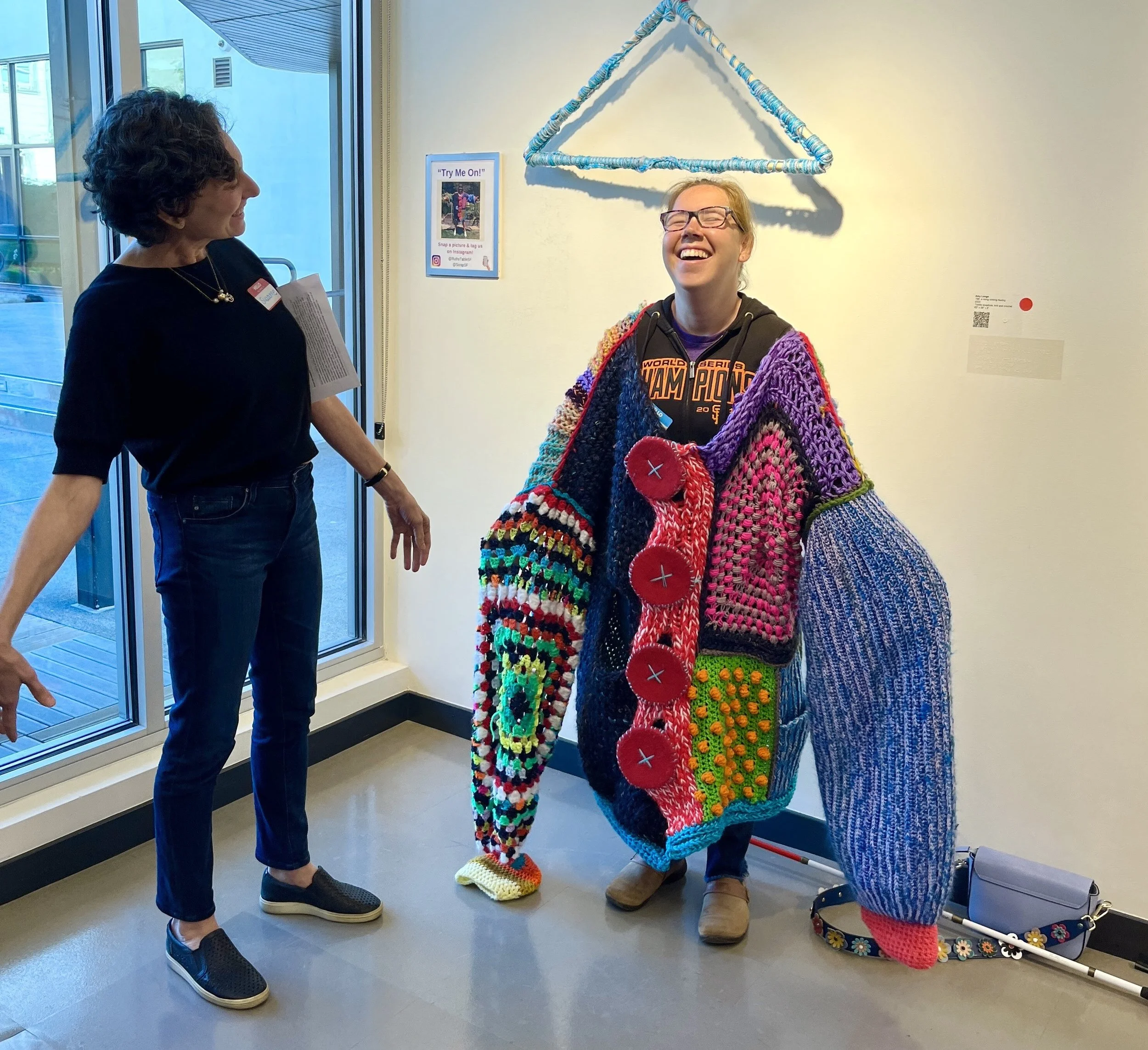 A woman and a girl smiling and talking inside a building near a wall with a woven art piece hanging above them. The woman is wearing a black shirt, blue jeans, and black slip-on shoes, holding a piece of paper. The girl is dressed in colorful, crocheted clothing, glasses, and smiling broadly.