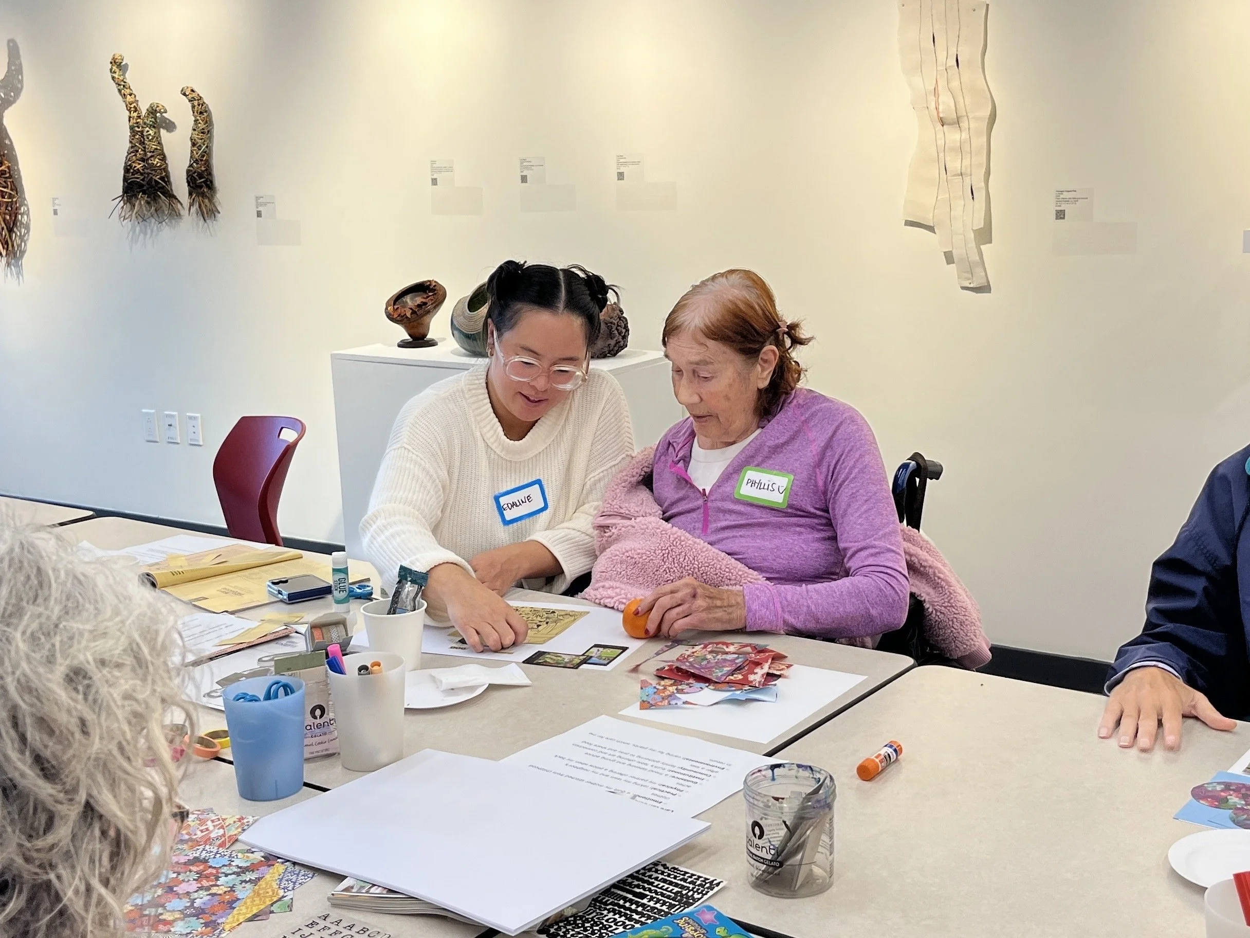 Two women sitting at a table engaging in arts and crafts activities. The woman on the left has dark hair styled in pigtails, glasses, and a name tag that says 'Eloise.' The woman on the right has red hair, is wearing a purple jacket, and has a name tag that says 'Philisc.' The table has various craft supplies, papers, and scissors, and there are artworks hanging on the wall behind them.