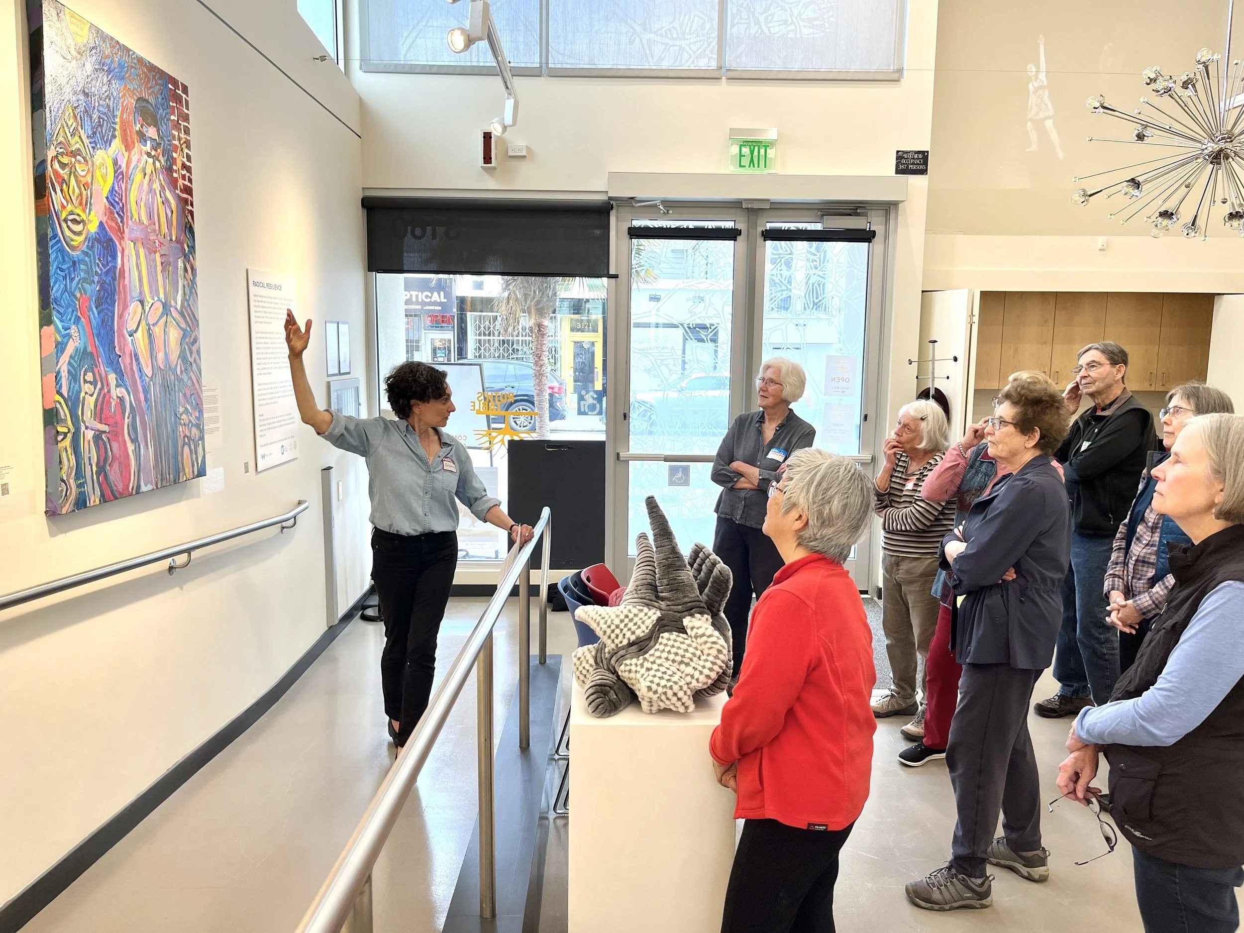 A group of people gathered inside an art gallery, listening to a woman who is gesturing towards a colorful painting on the wall. The gallery has large windows and modern lighting fixtures.