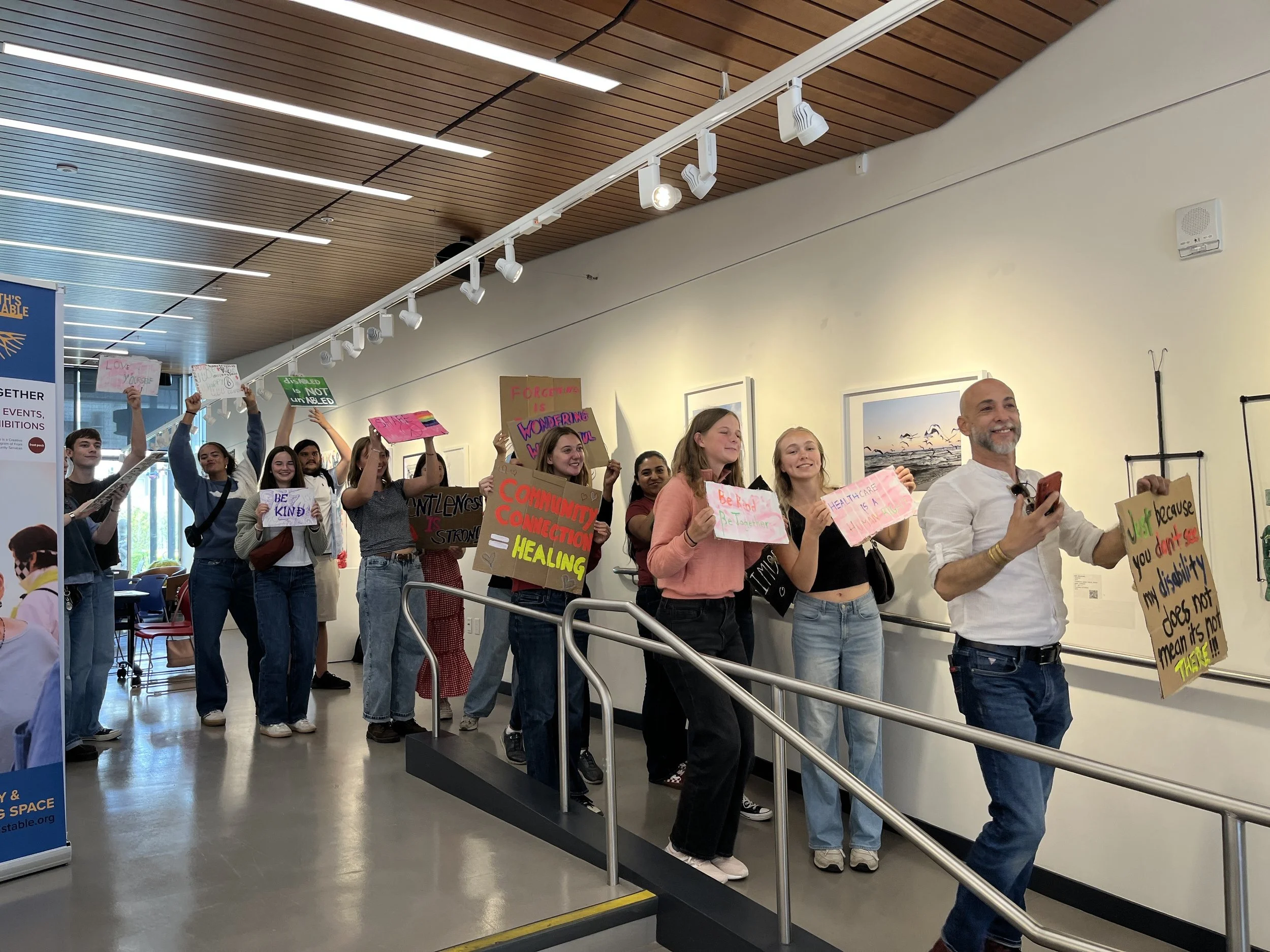 A diverse group of people, including young adults and adults, holding signs and banners with messages about community, health, and disability awareness, standing inside a modern room with wooden ceiling and track lighting.