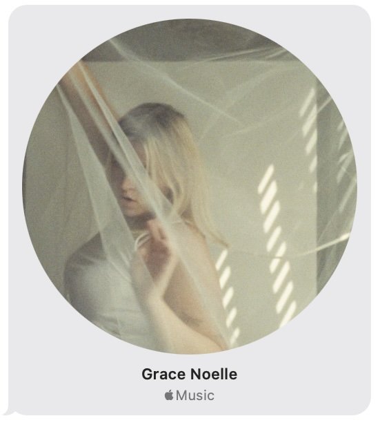A woman with blonde hair behind a semi-transparent curtain with sunlight streaming through blinds.