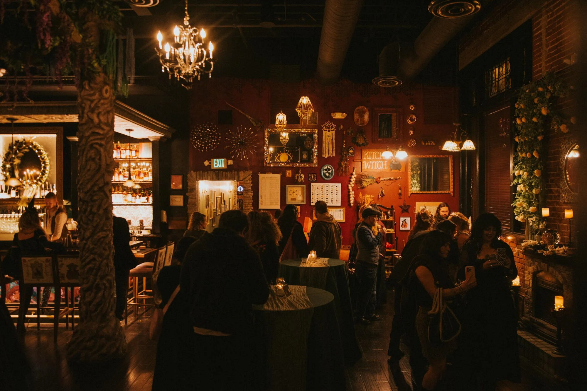 A dimly lit restaurant or bar with hanging chandeliers and warm lighting. Decor includes various framed pictures, plants, and a sign that reads 'KITCHEN WITCH.' There are multiple groups of people seated at tables, some using their phones, creating a lively atmosphere.