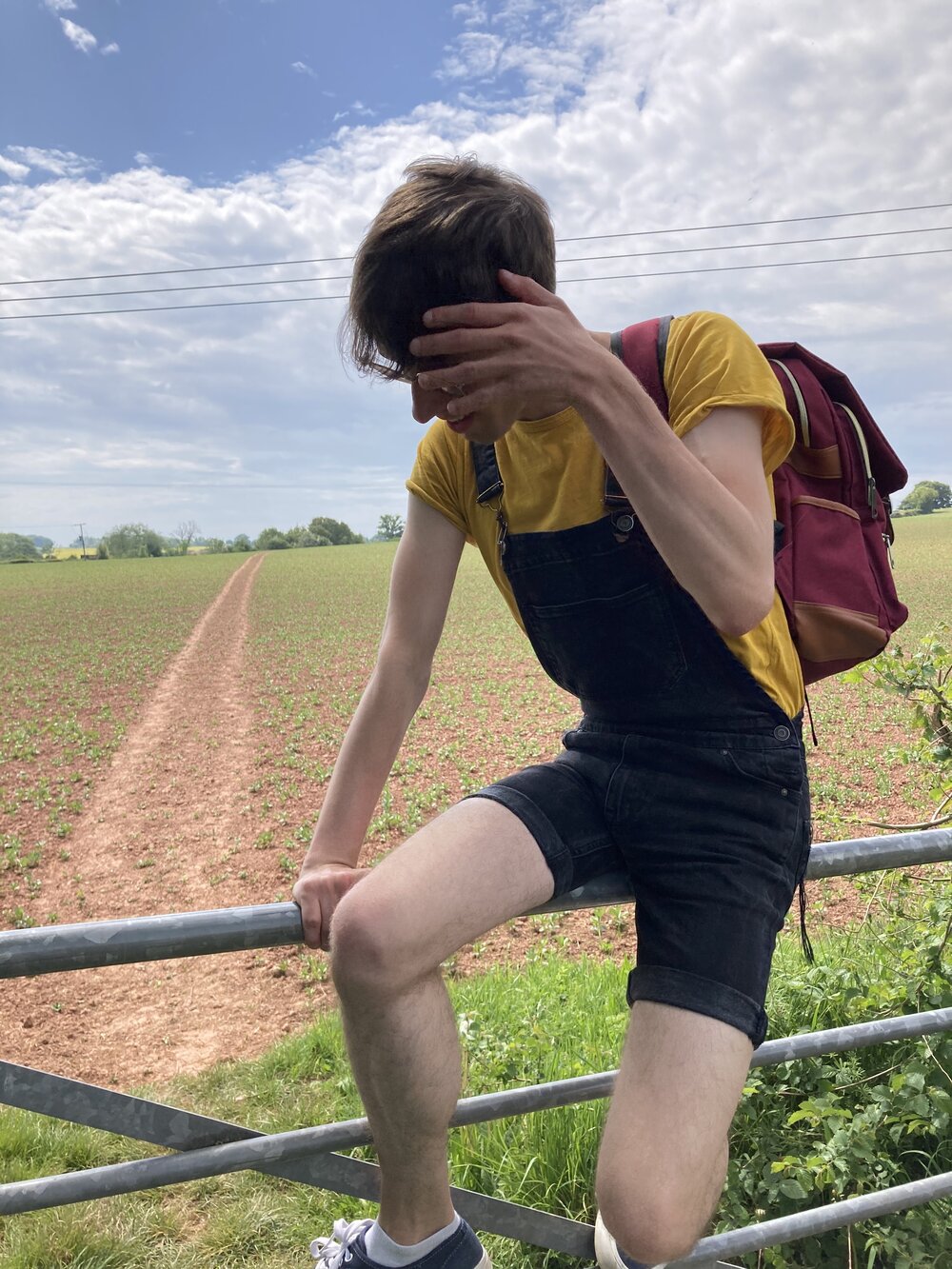 Dec pictured resting on a fence in the countryside. He's wearing a shortsleeved yellow t-shirt and fitted black short dungarees. Photo: Declan Bowring/Lacuna Voices 