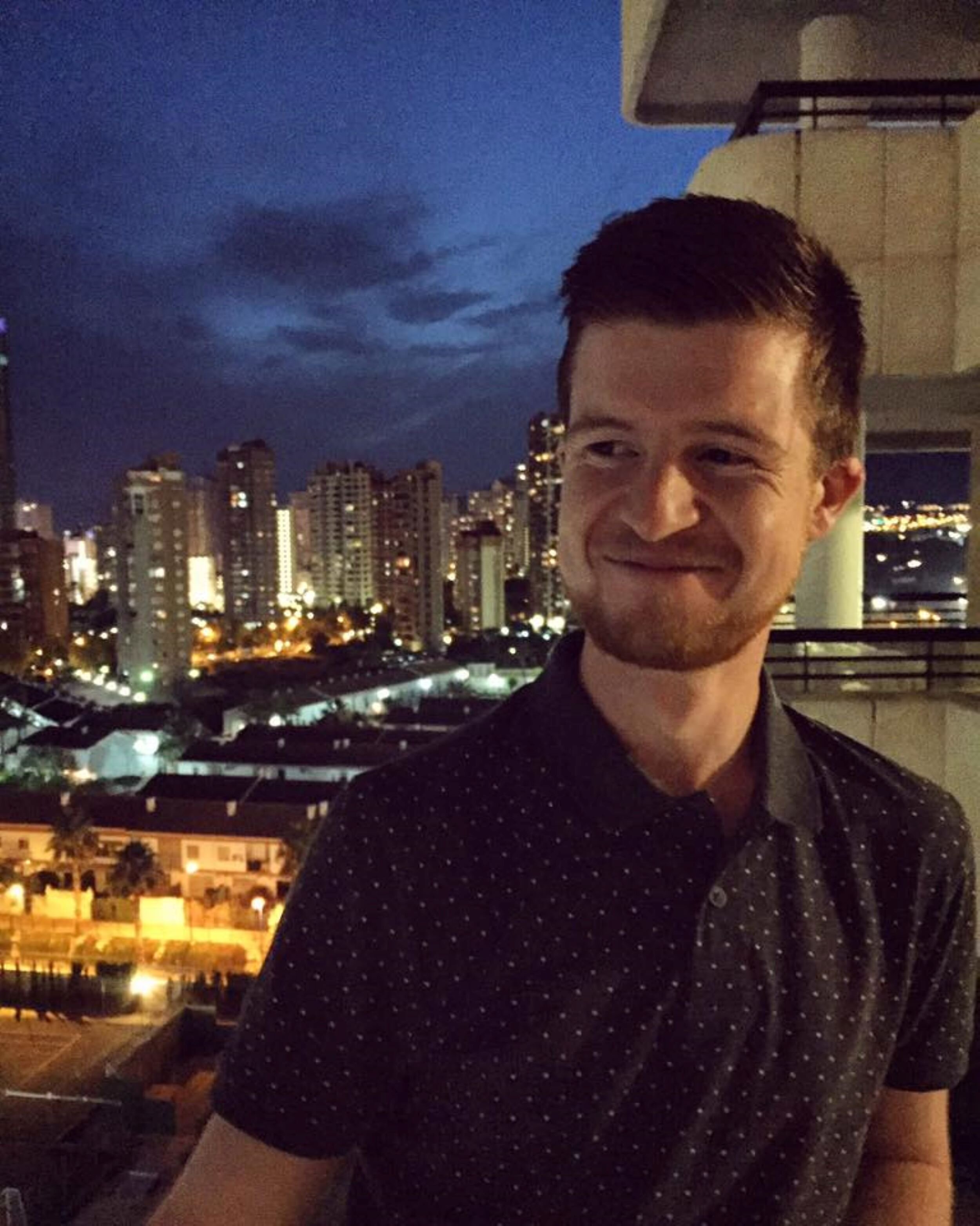 Alex enjoying the sites in Spain 2016. He's pictured wearing a short sleeve black button up shirt, with the metropolitan night skyline lit up behind him. Photo: Alex Waite