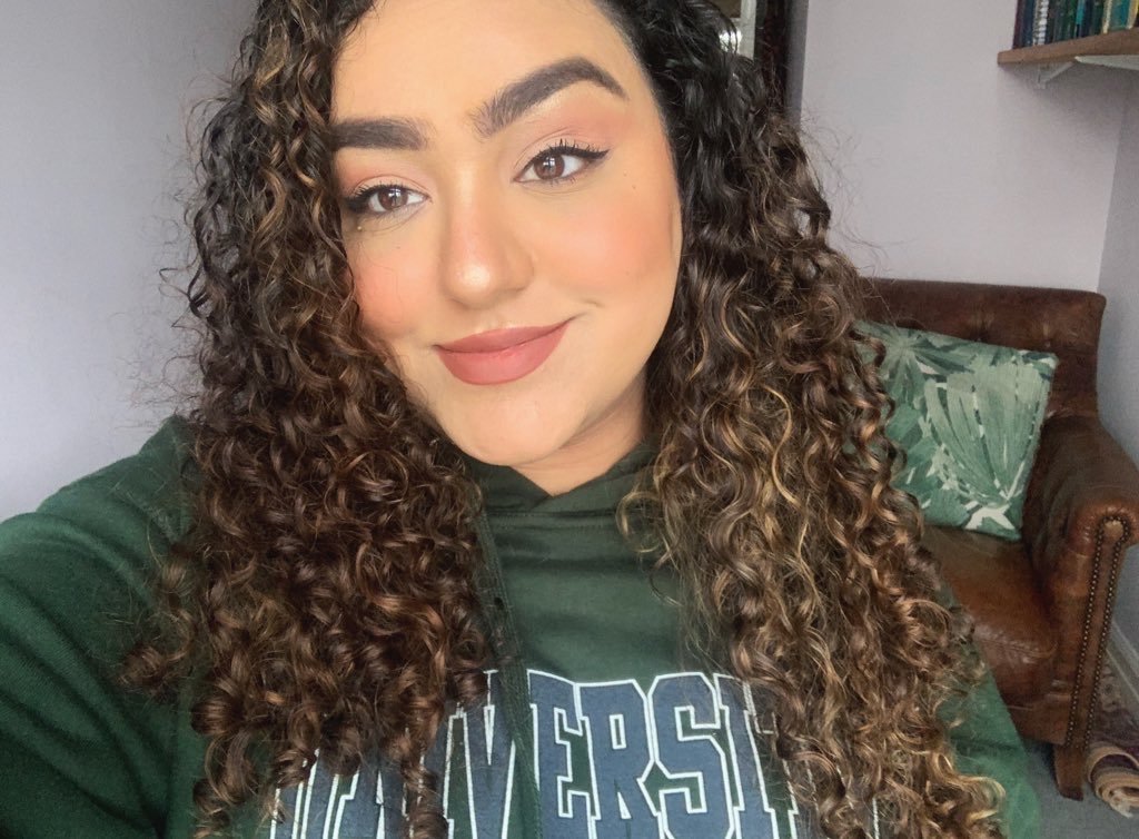 A selfie of Sarah Harris now. Her brunette hair is curly and she is wearing a green university hoodie | UK