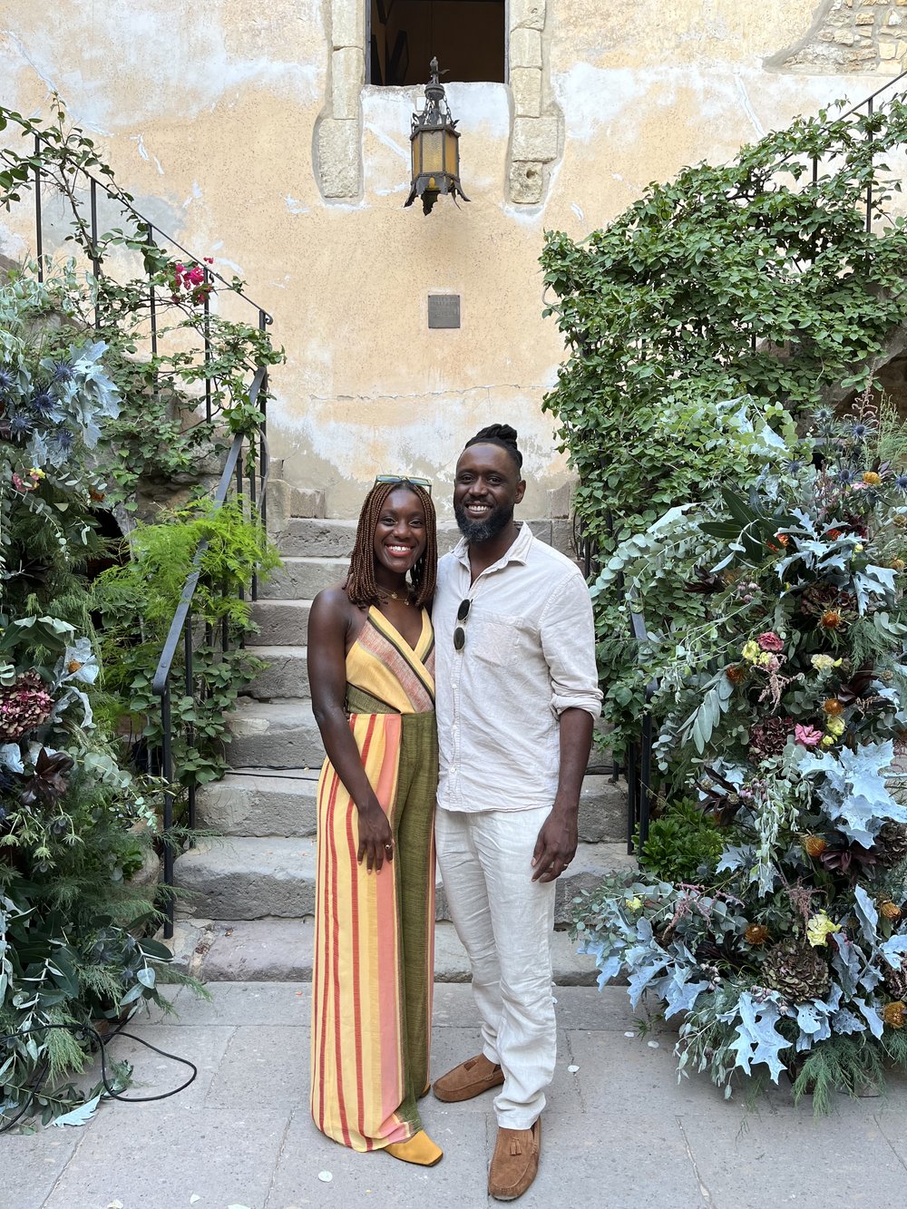 Shakira and Patrick on their honeymoon. She is wearing a beautiful strappy and cutout jumpsuit. He is wearing a white linen set and they stand by a staircase covered in green foliage. Photo Shakira Peronet.jpg