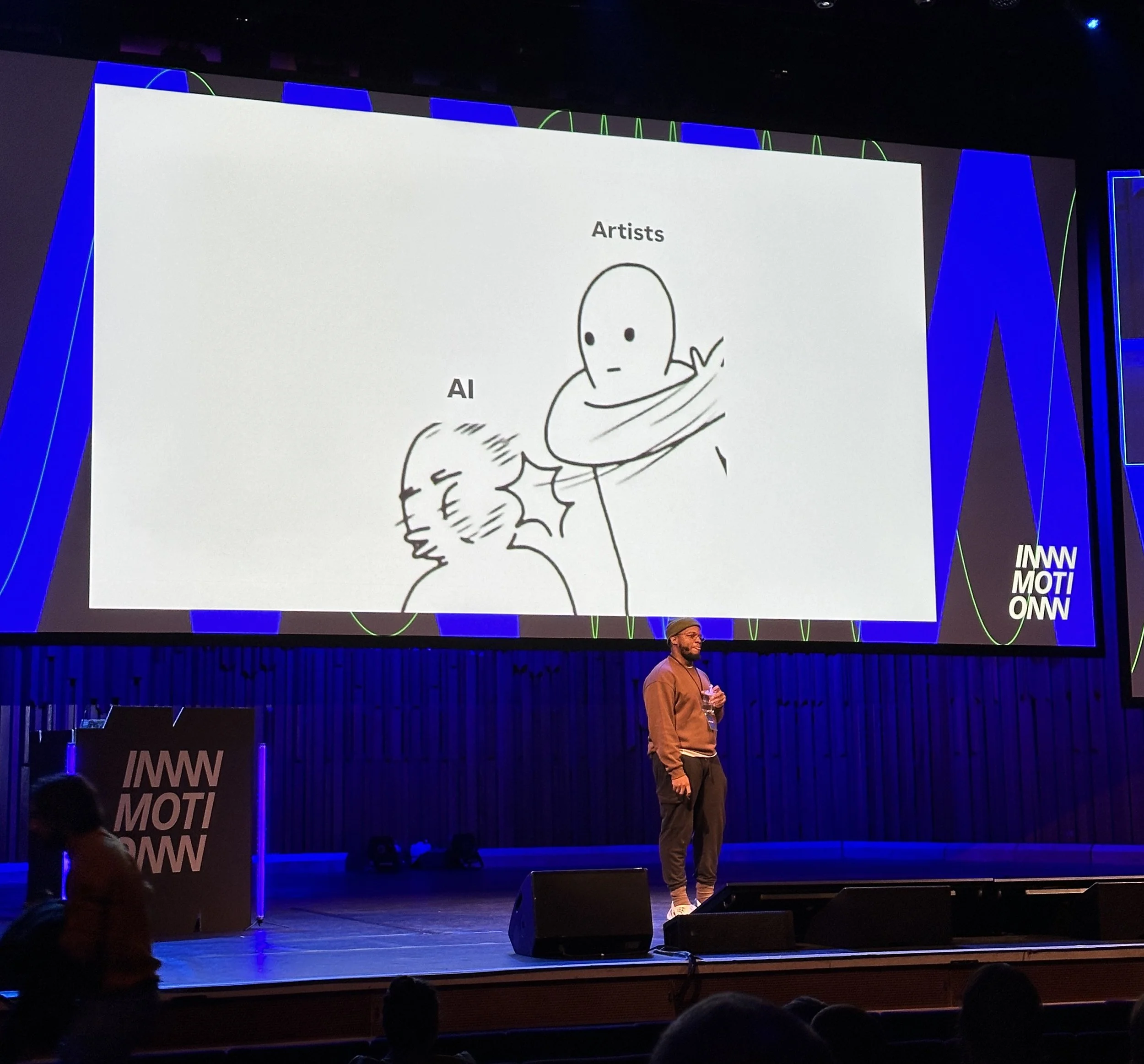 Photo from Maxime Manga's talk at InMotion 2025. He is on stage and the slide behind him shows a drawing of two simple characters: one labelled "Artists" is slapping the one labelled "AI" around the face.