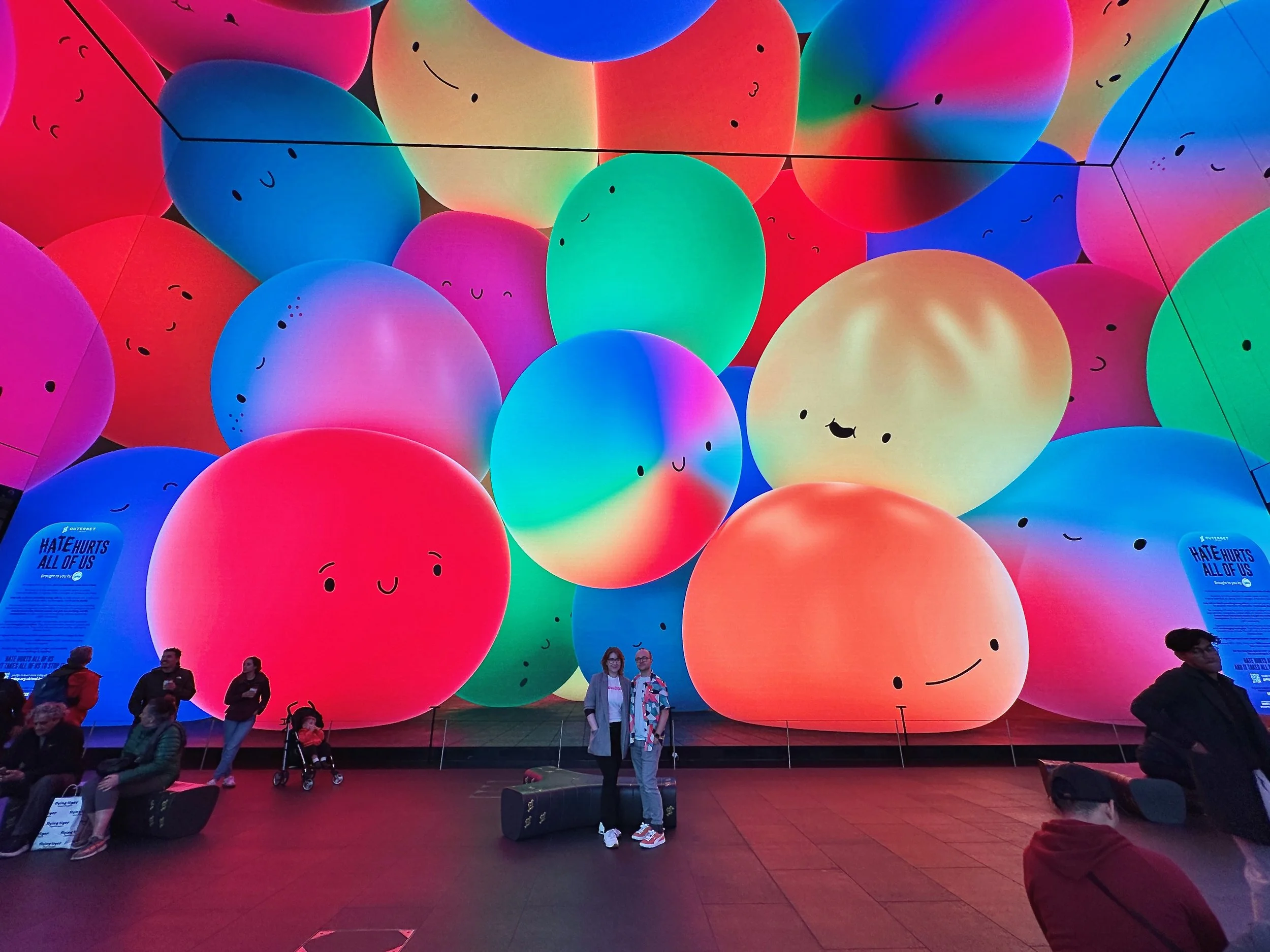 A photo of Chicken Fruit's Jonny and Lindsey at the Outernet. Behind them, on the giant screen of the south wall, are many colourful squidgy characters: Galop's Squidgies.