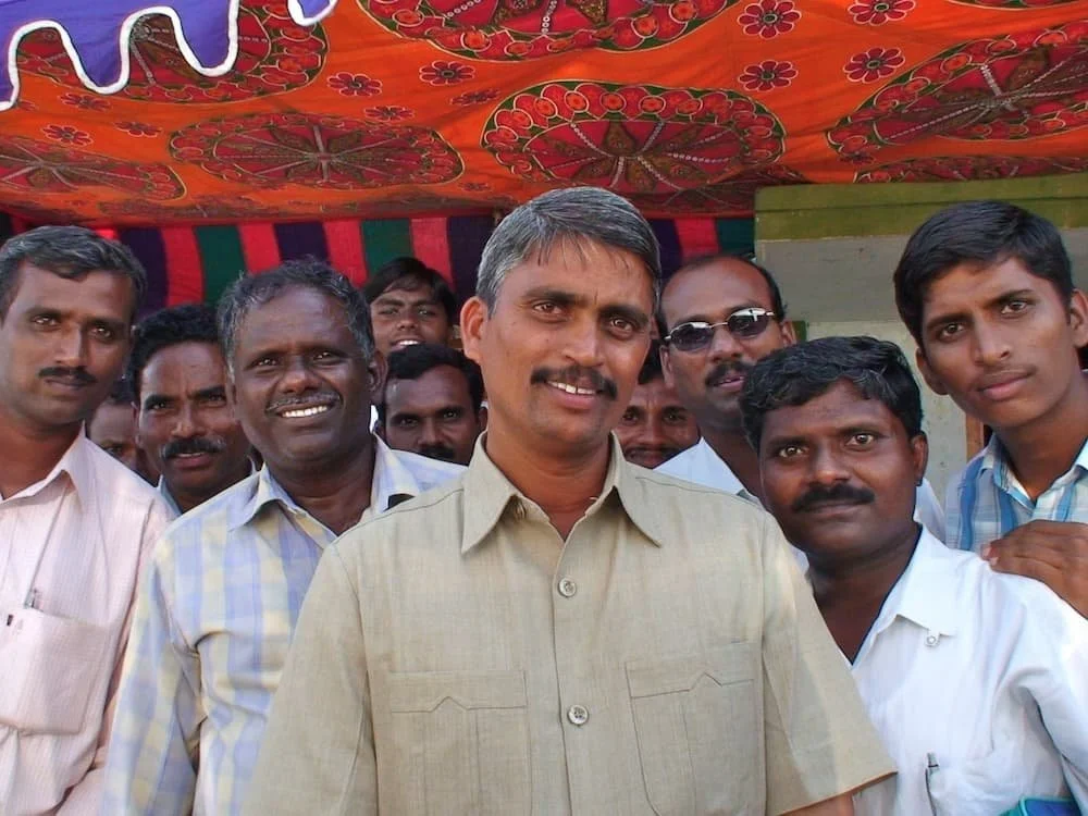 Un groupe d'hommes indiens, certains souriants, d'autres sérieux, debout ensemble à l'extérieur, sous un auvent coloré.